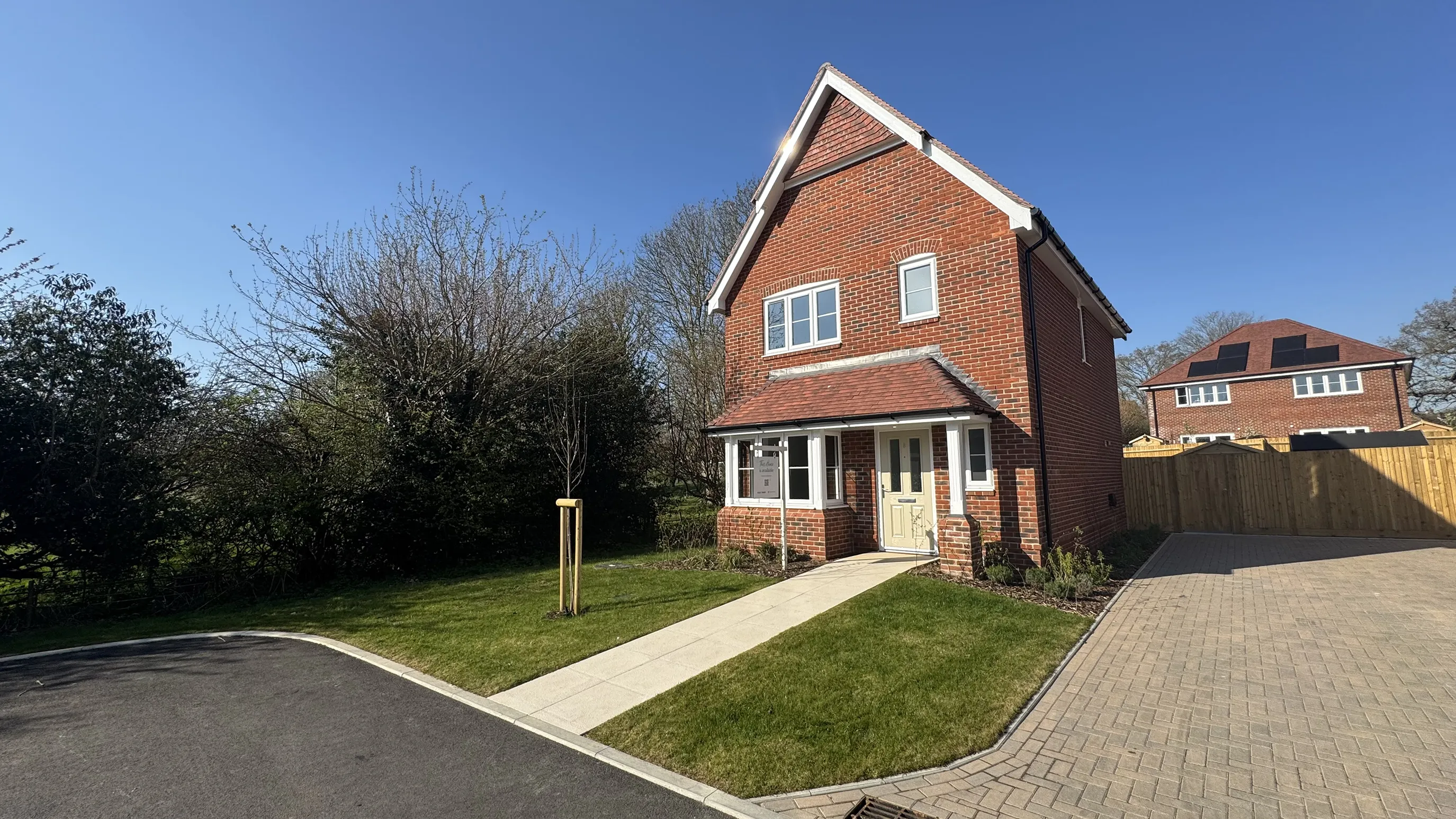 Two-story brick house with a triangular roof, a small front yard, and a driveway on the right. A clear blue sky and a similar house are visible in the background.
