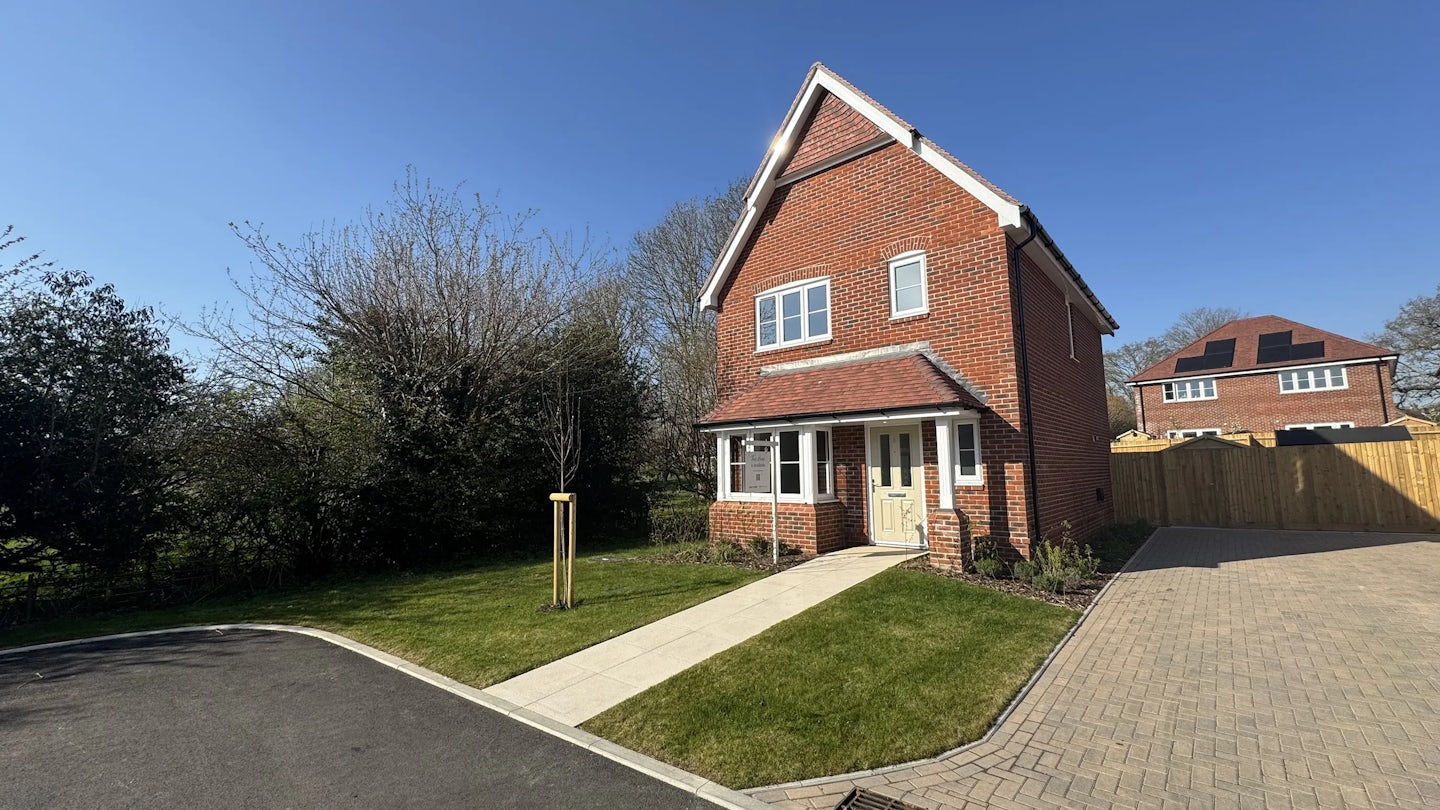 Two-story brick house with a triangular roof, a small front yard, and a driveway on the right. A clear blue sky and a similar house are visible in the background.