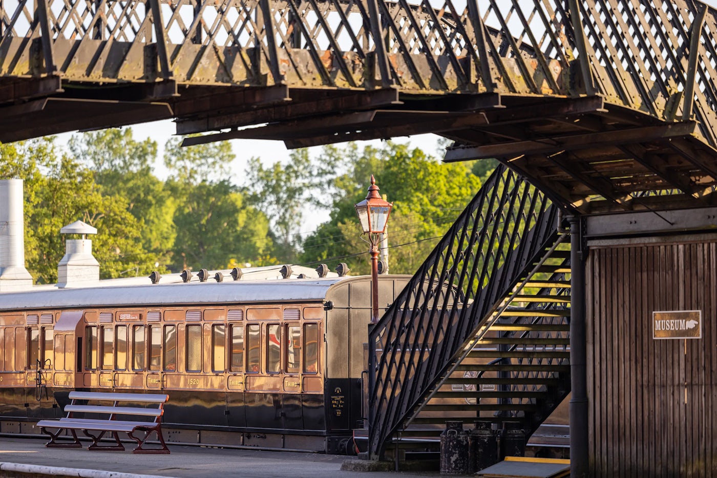 Vintage train station scene with an old-fashioned train car, wooden bench, staircase to an overhead pedestrian bridge, lamppost, and a 'MUSEUM' sign on a wooden structure, with trees and another building in the background.