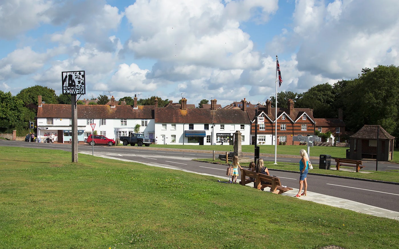 Picturesque village scene with traditional English houses, a road with parked cars, grassy area with benches and people, and a Union flag on a flagpole.