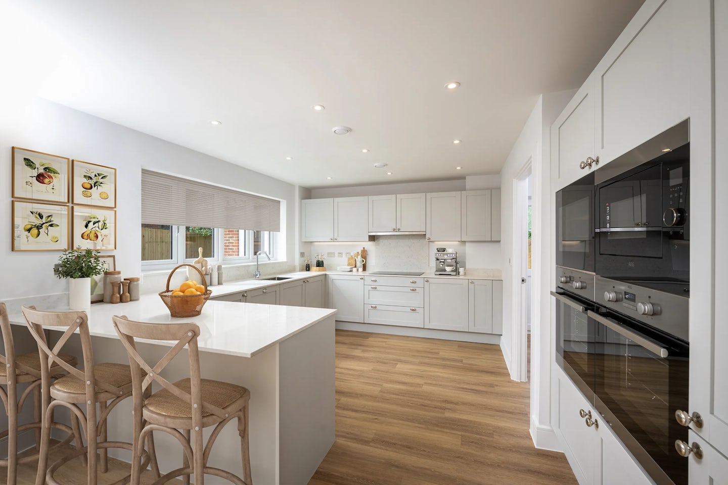 Modern kitchen with white cabinets, wooden flooring, and a central island with three wooden chairs and a basket of oranges. Built-in oven and microwave on the right, framed fruit pictures and countertop with plants and utensils on the left, with natural light coming through partially drawn blinds.