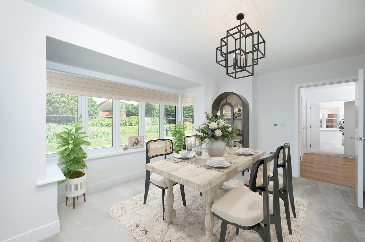 Modern dining room with a wooden table set for four, featuring plates, bowls, cutlery, and a floral centerpiece. Natural light enters through a large window with beige blinds, flanked by two potted plants. A geometric black chandelier hangs above, with an arched shelf unit and an adjacent room visible in the background.