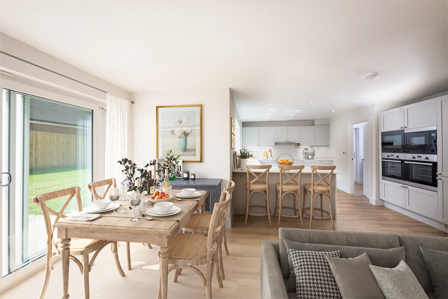 Modern open-concept kitchen and dining area with a wooden dining table set for four, decorated with flowers and oranges. A kitchen island with four bar stools and light-colored cabinets is in the background, with built-in appliances on the right. A grey sofa is partially visible in the foreground.