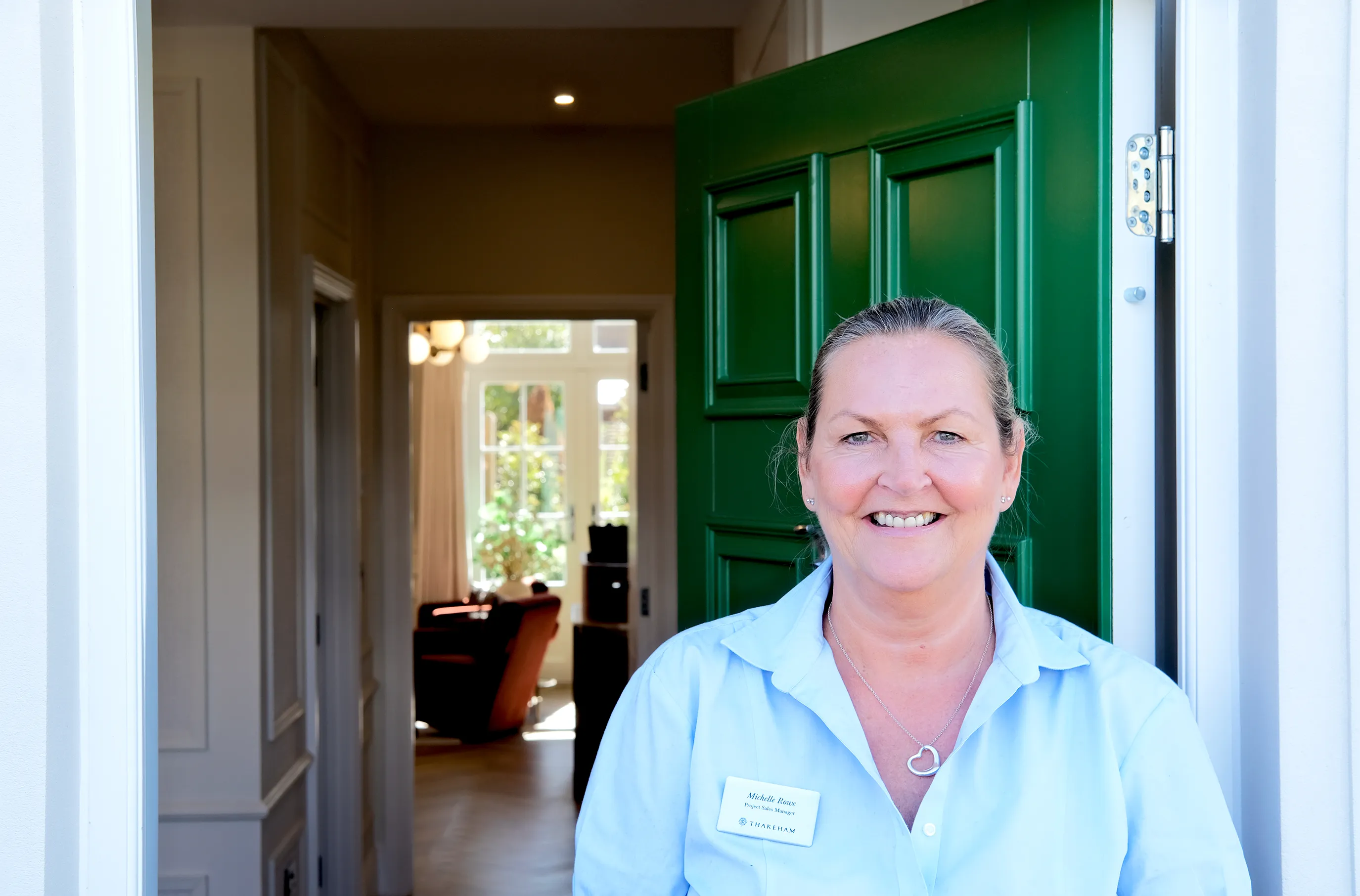 Michelle wears a blue shirt and stands in front of a green door. She has blond hair tied back.
