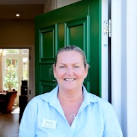 Michelle wears a blue shirt and stands in front of a green door. She has blond hair tied back.