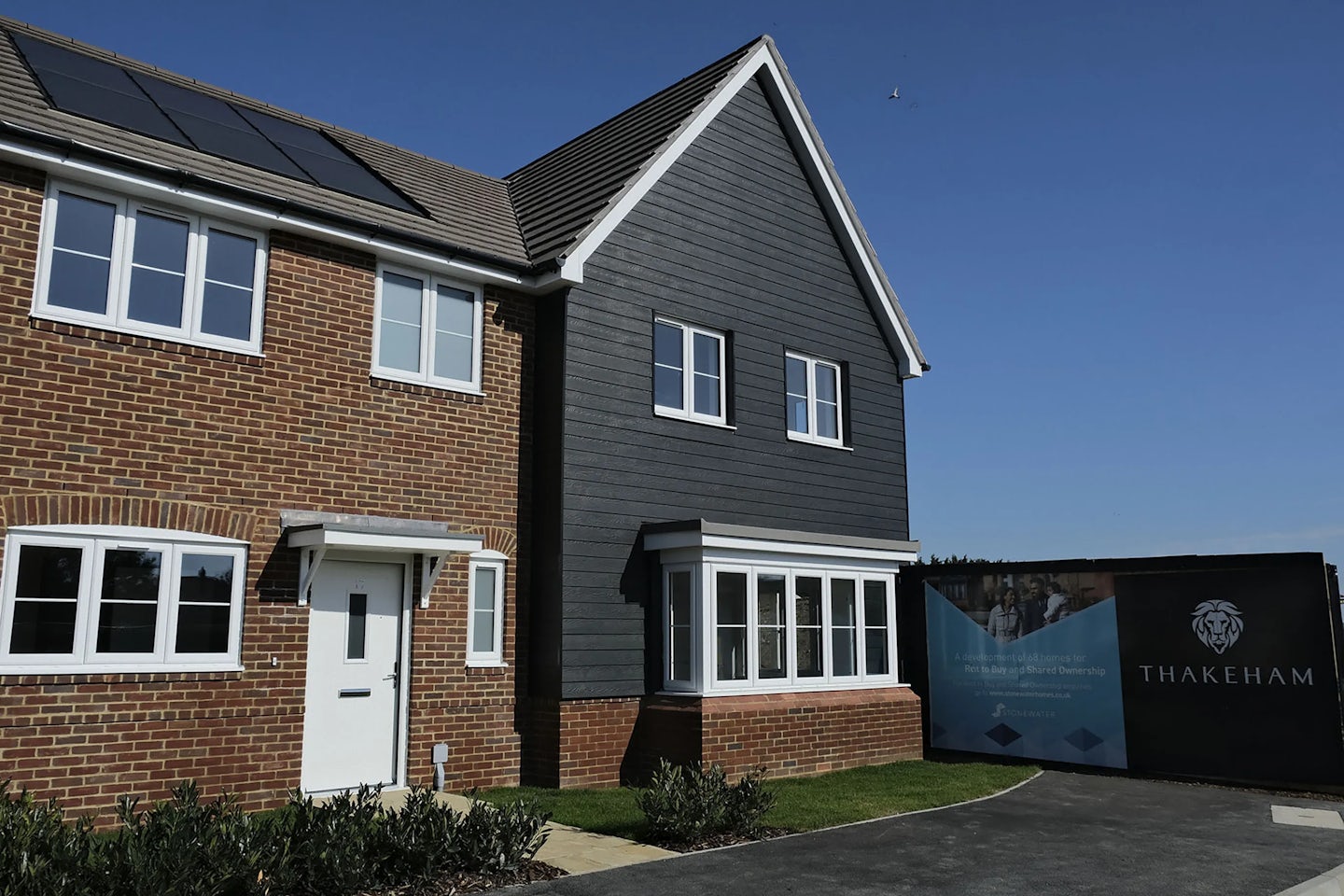 Modern two-story house with red brick and dark grey siding, featuring solar panels on the roof and a 'THAKEHAM' sign in front, under a clear blue sky.