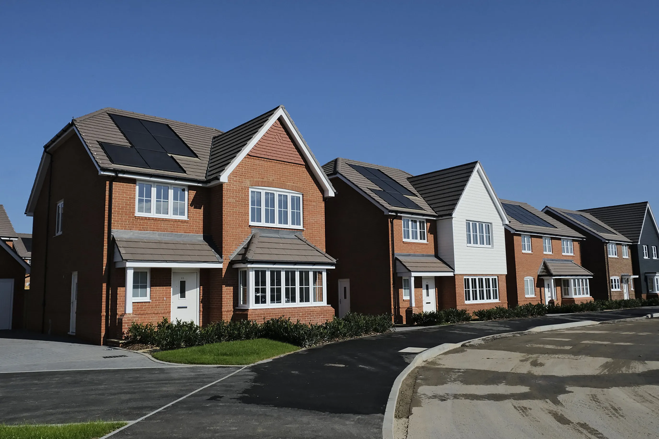 Row of modern detached brick houses with white accents and solar panels on the roofs, set along a paved suburban street under a clear blue sky.