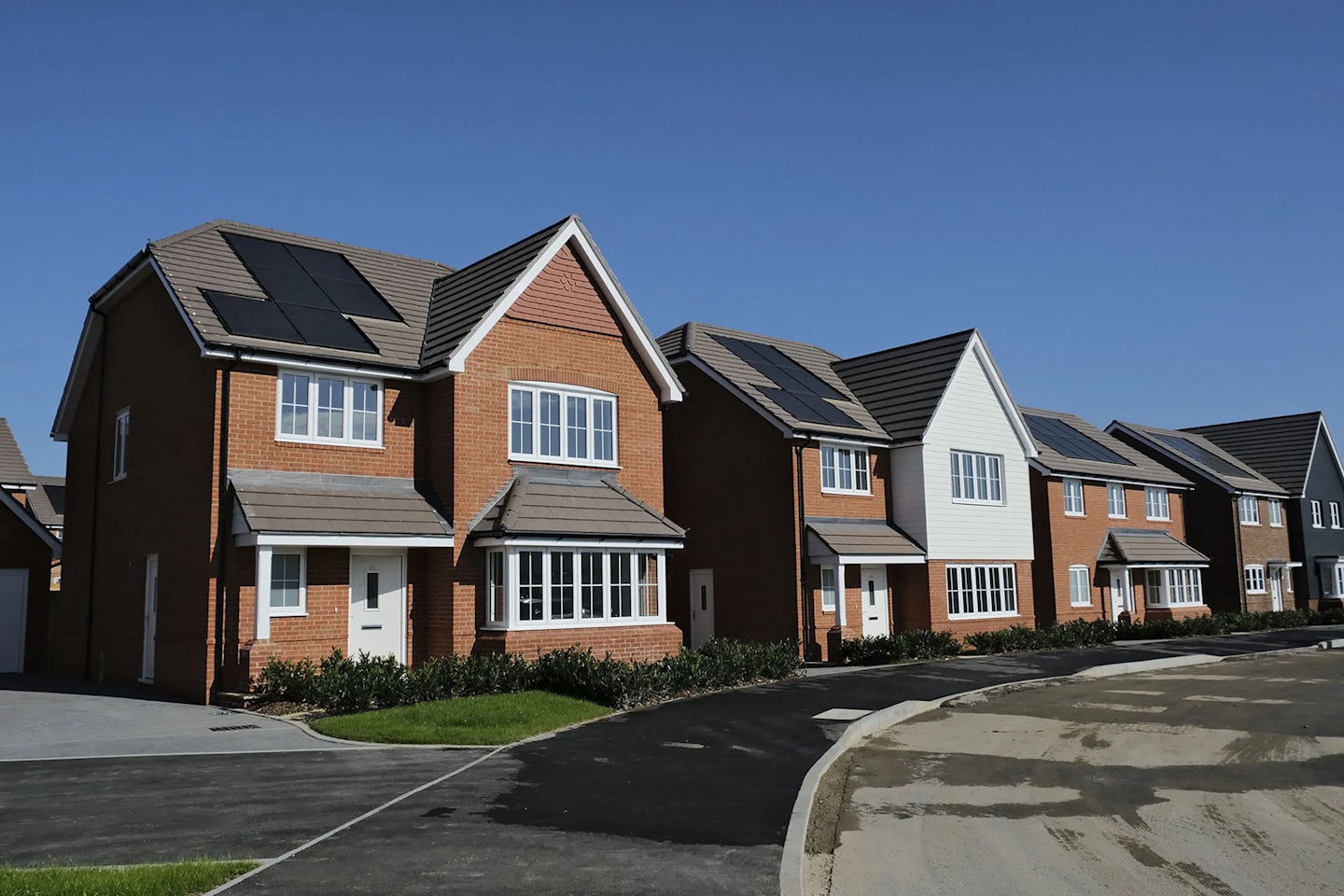 Row of modern detached brick houses with white accents and solar panels on the roofs, set along a paved suburban street under a clear blue sky.