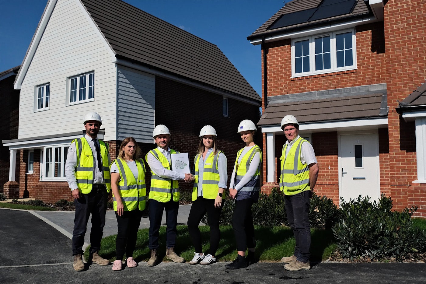 Six individuals in high-visibility vests and hard hats pose in front of two newly built houses, one featuring solar panels, marking a construction milestone.