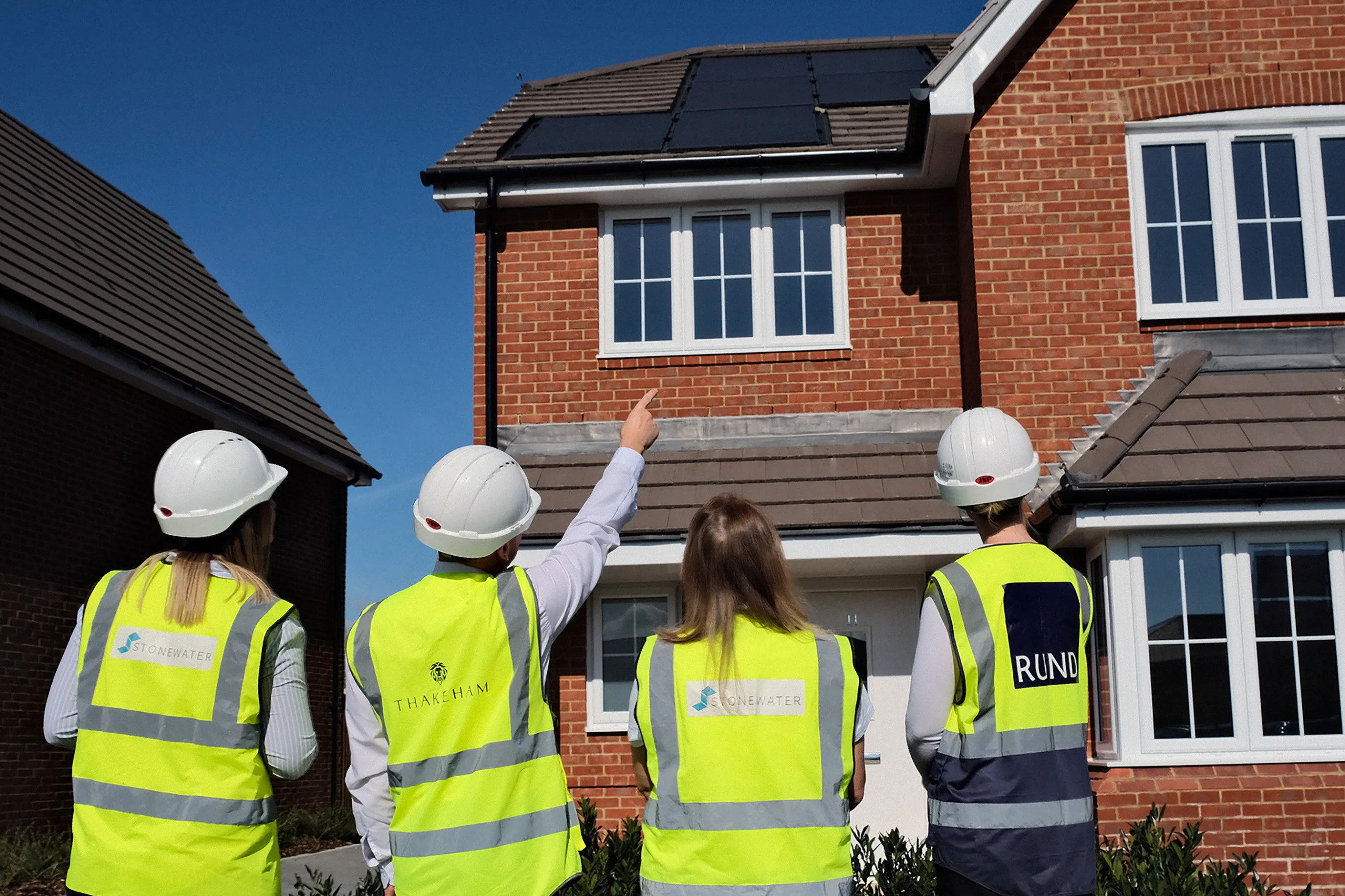 Four individuals wearing safety helmets and high-visibility vests stand in front of a brick house with solar panels on the roof, appearing to inspect or discuss the installation.
