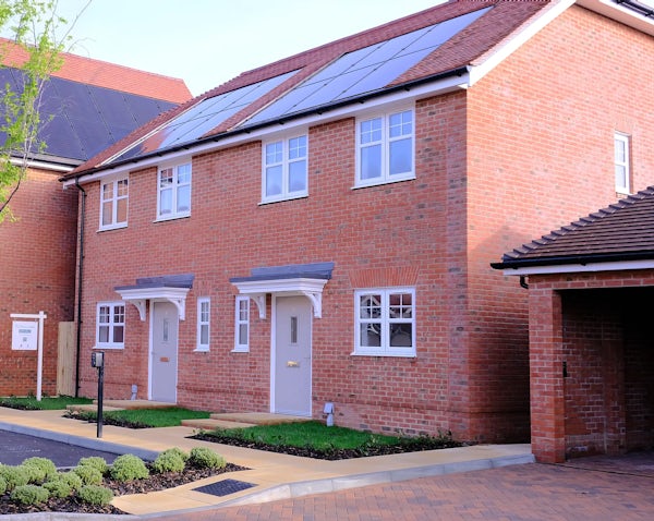 Modern brick houses in a residential area, one with a grey car parked in front and a bay window, the other featuring solar panels and twin white doors with small porches. A small tree, trimmed bushes, and paved pathways are visible in the foreground.