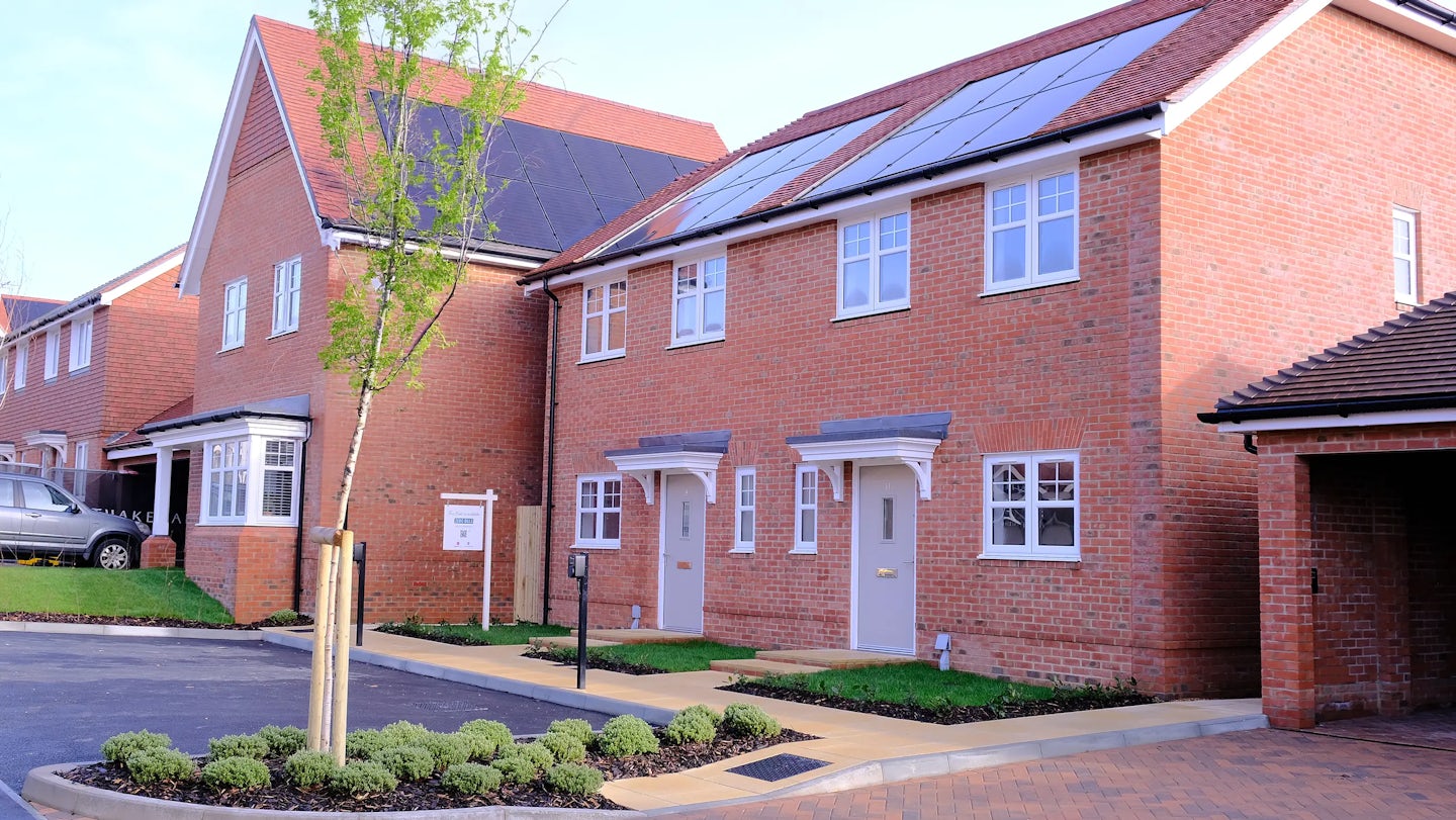 Modern brick houses in a residential area, one with a grey car parked in front and a bay window, the other featuring solar panels and twin white doors with small porches. A small tree, trimmed bushes, and paved pathways are visible in the foreground.