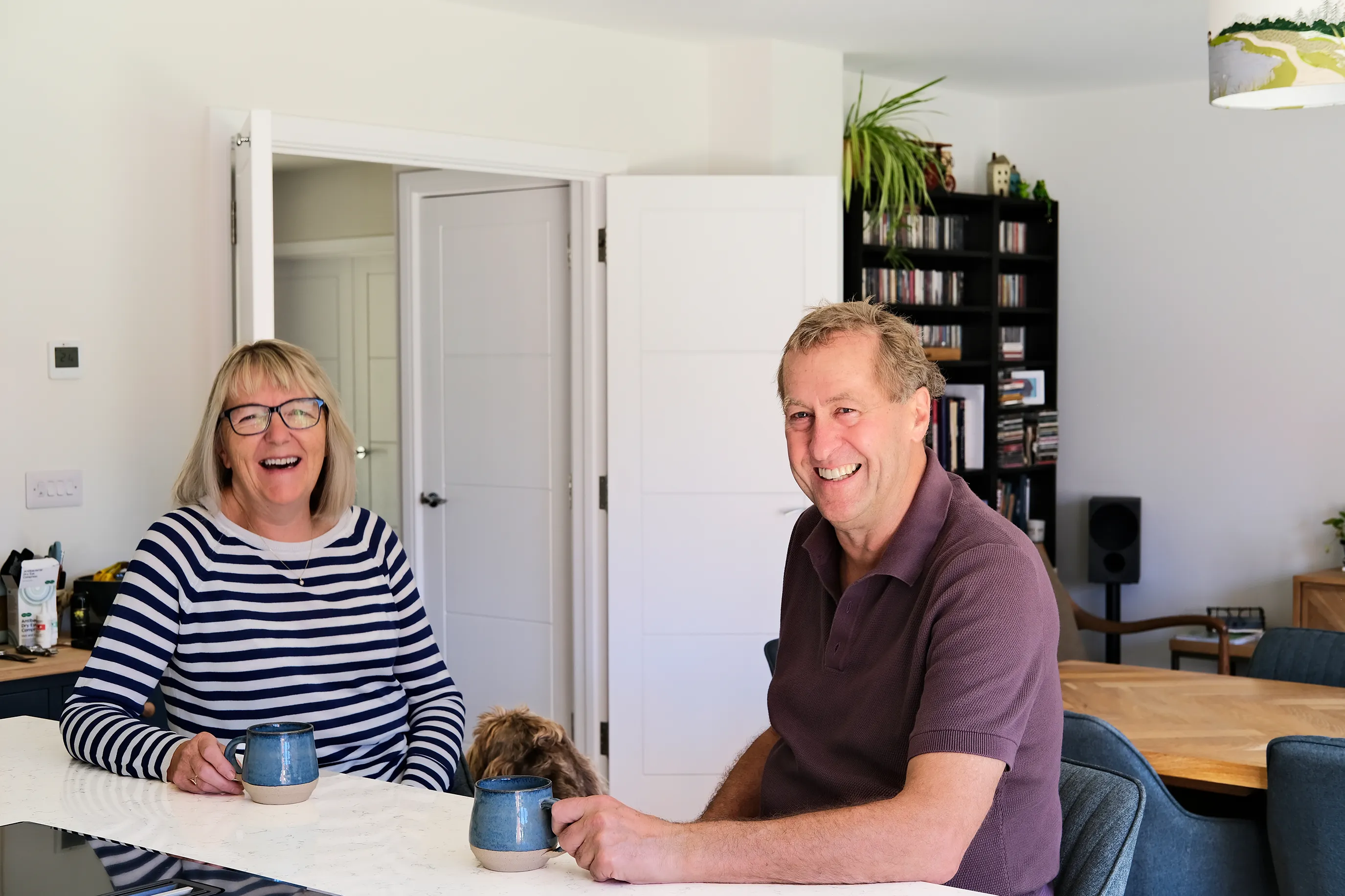 Two people sitting at a dining table in a modern, well-lit room, each holding a ceramic mug. A small dog is partially visible between them. Behind them is an open white door, a black shelving unit with books and speakers, and a wooden dining table with chairs.