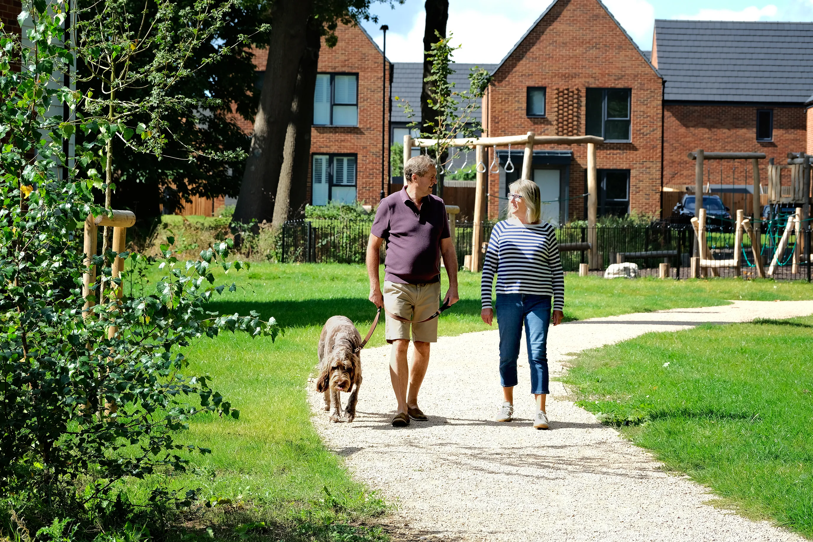 Two people walking a dog along a gravel path in a green residential area with trees, grass, and modern brick houses in the background. A small playground structure is visible behind them.