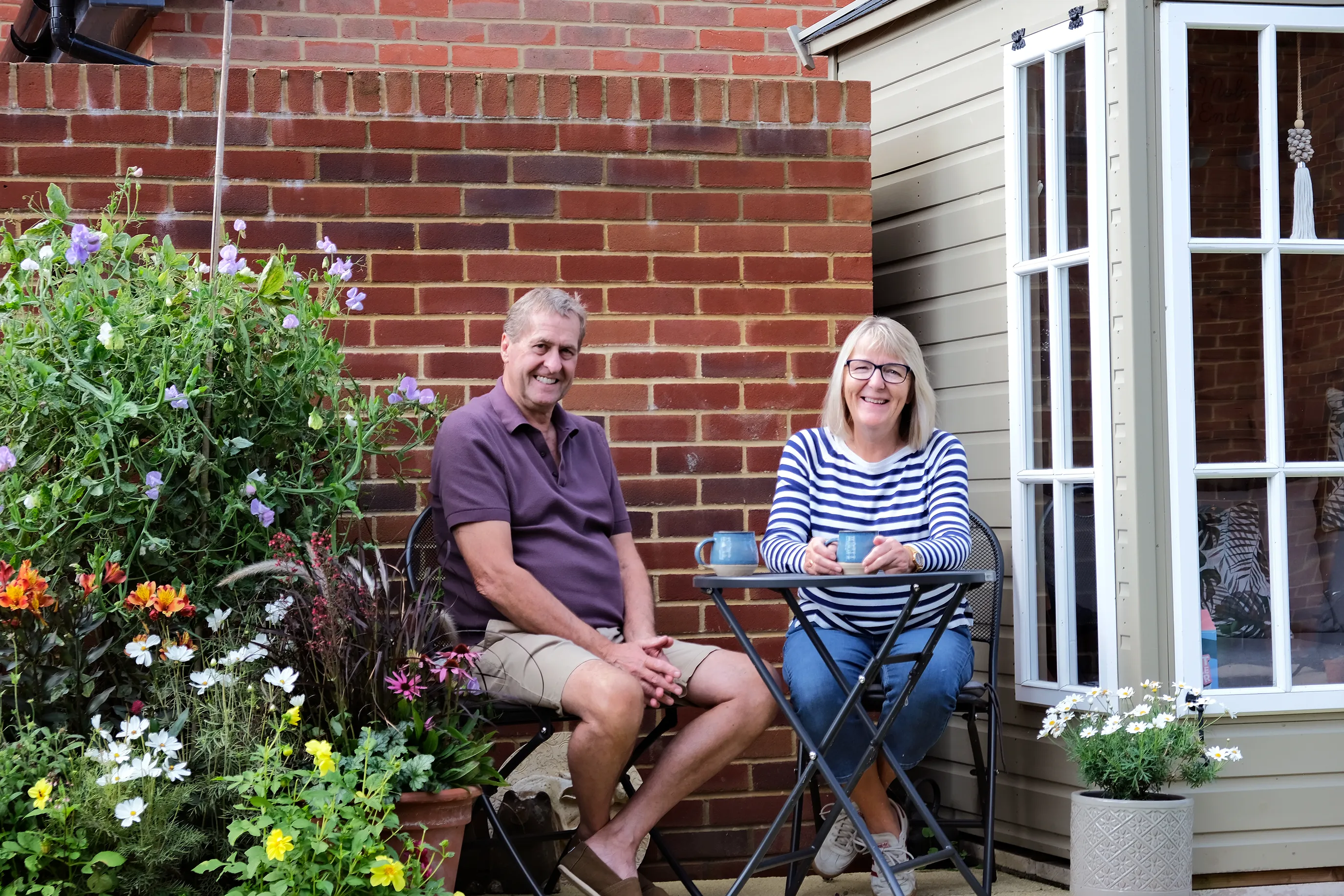 Two people sitting at a small outdoor table in a garden area, surrounded by colorful flowers and plants, with a brick wall and a house window in the background. Both have mugs on the table.