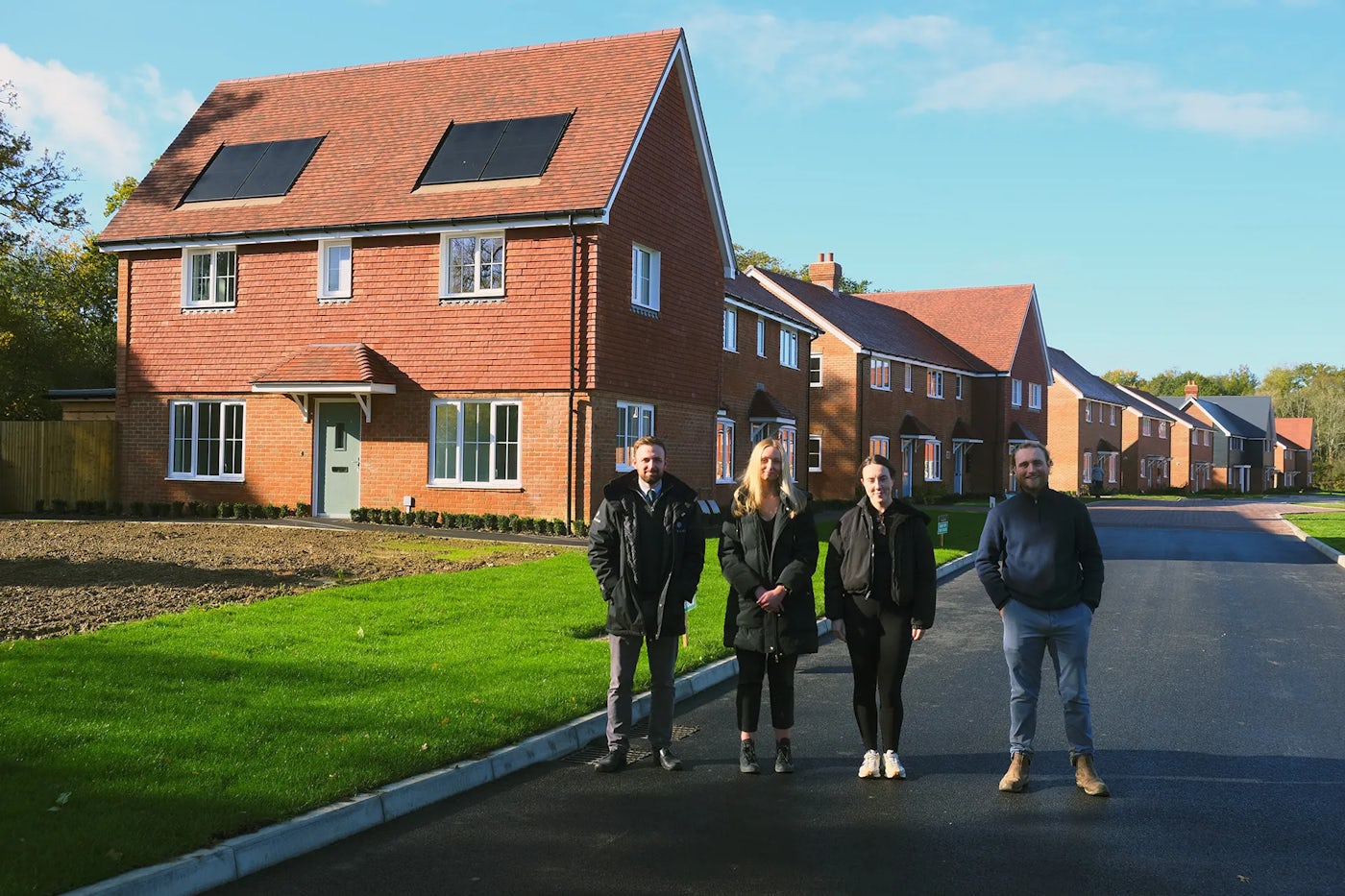 Four people standing on a newly paved street in front of a row of modern red-brick houses with pitched roofs and solar panels. The houses have white-framed windows and small front gardens, and the area appears to be a new residential development under clear blue skies.
