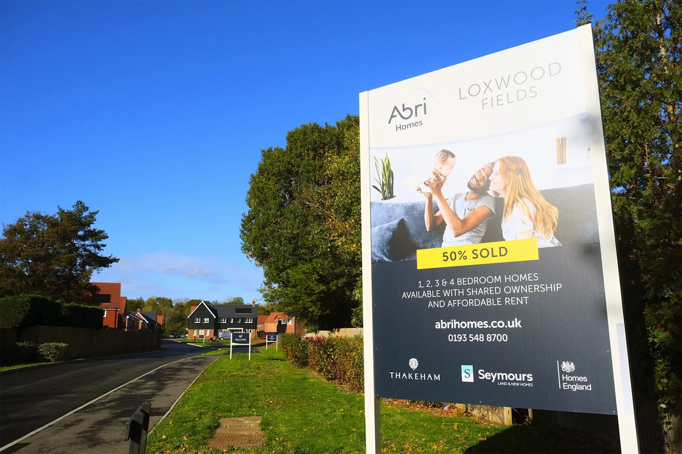 Large roadside sign for Abri Homes at Loxwood Fields advertising 1, 2, 3, and 4-bedroom homes with shared ownership and affordable rent. The sign shows a family image and includes text stating ‘50% SOLD’ along with contact details and logos for Thakeham, Seymours, and Homes England. Behind the sign is a quiet residential street with houses and trees under a bright blue sky.