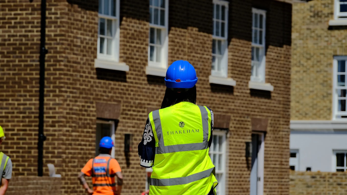 Construction site scene showing a person wearing a bright yellow Thakeham-branded safety vest and a blue hard hat, standing in front of a brick building with multiple white-framed windows. Other workers in safety gear and hard hats are visible in the background.