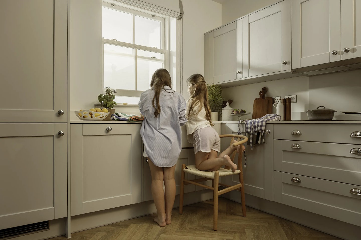 Two children in a bright kitchen looking out of a large window. One child is standing while the other kneels on a wooden chair. The kitchen features light-colored cabinets, a wooden herringbone floor, and countertops with items including a bowl of lemons, a folded tea towel, a potted plant, and a wooden chopping board.