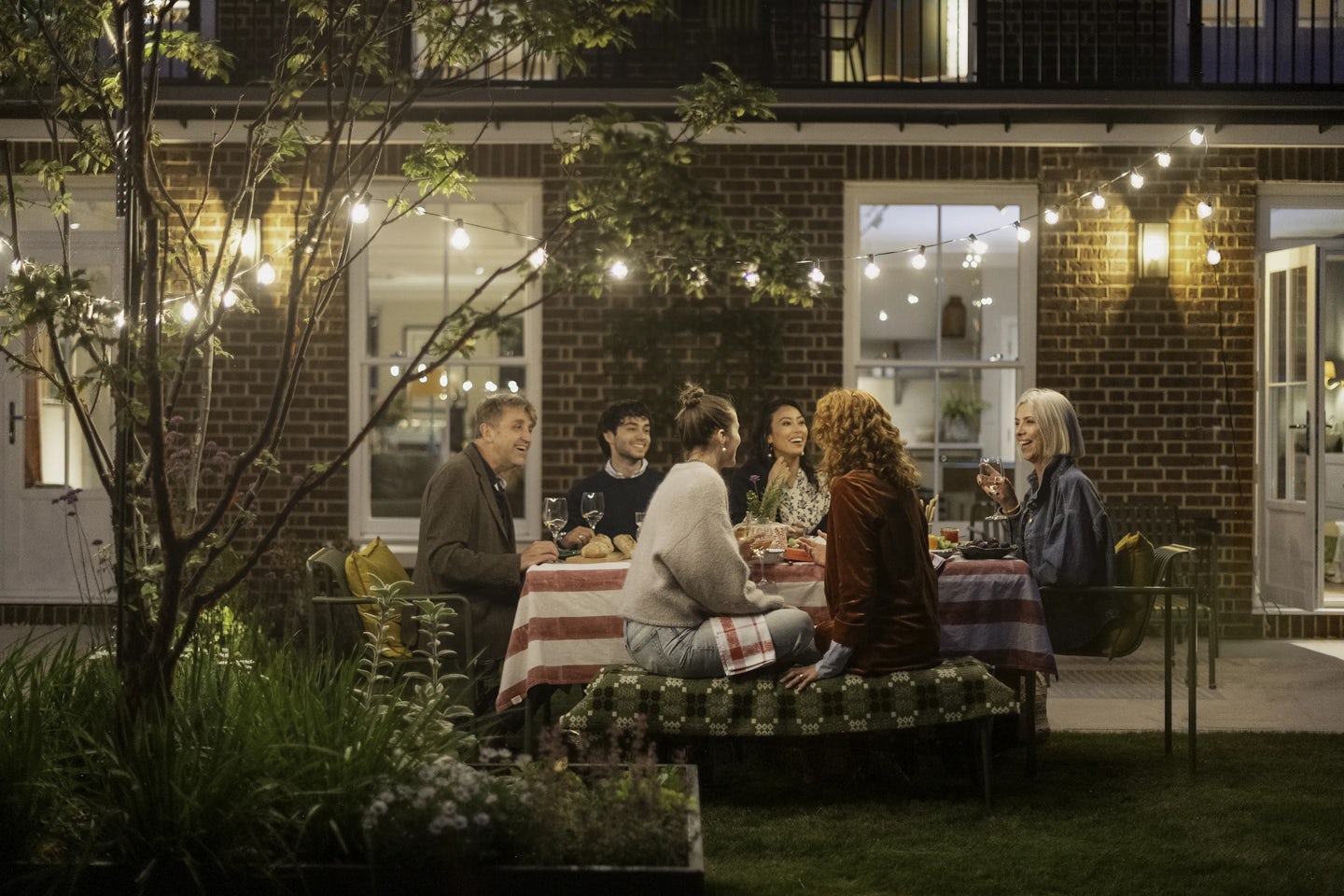 Outdoor evening gathering in a garden with six people seated around a rectangular table covered with a red-and-white striped tablecloth. The table is set with plates, glasses, and bottles, and string lights are hanging above, illuminating the scene. Behind the group is a brick house with large windows and a door leading to the patio.