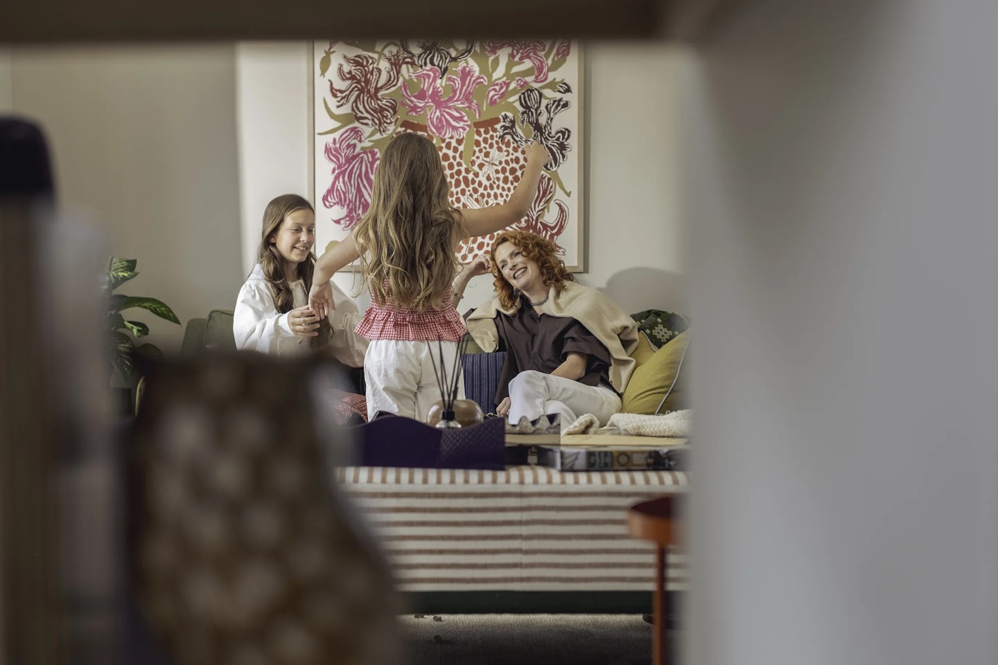 Three people in a living room, with one standing and raising an arm while two others sit on a sofa. The sofa has cushions and a blanket, and a large floral artwork hangs on the wall behind them. Indoor plants are placed near the seating area, and the foreground shows part of a striped piece of furniture.
