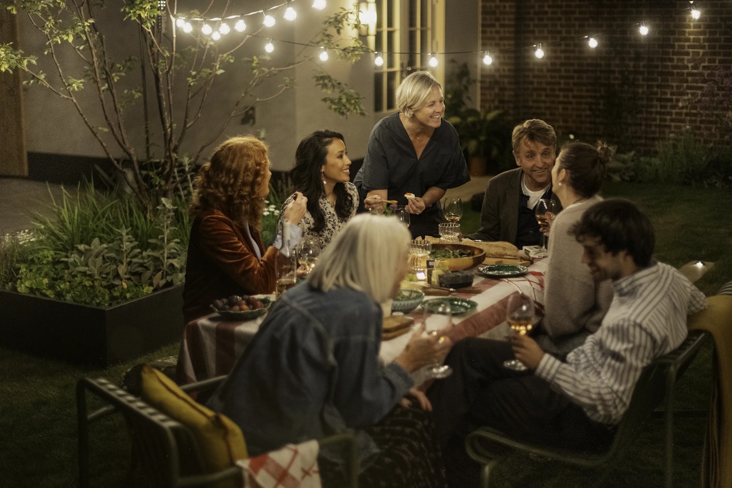 Group of people seated around a rectangular outdoor dining table in a garden at night. The table is covered with a red-and-white checkered cloth and set with plates, glasses of wine, and bowls of food. String lights hang overhead, illuminating the scene. The background shows a brick wall, a door, and plants in raised beds.
