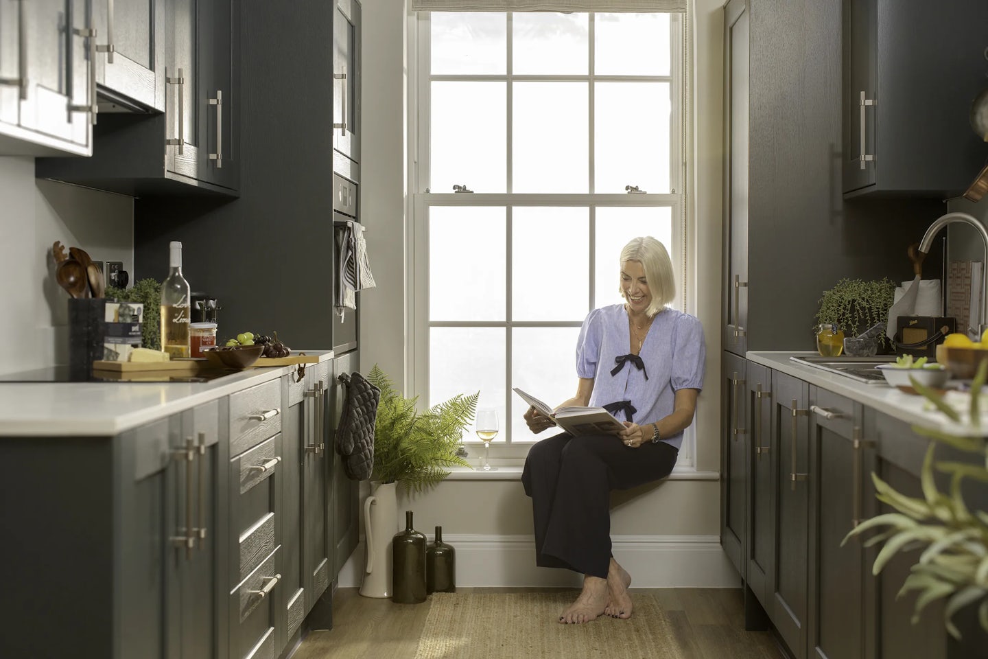 Person sitting on a wide windowsill in a modern kitchen, reading a book. The kitchen has dark cabinets, light countertops, and a stainless steel sink. On the counter are bottles, a cutting board with food items, and a potted plant. Two decorative bottles and a wine glass are placed near the window, which lets in bright natural light.