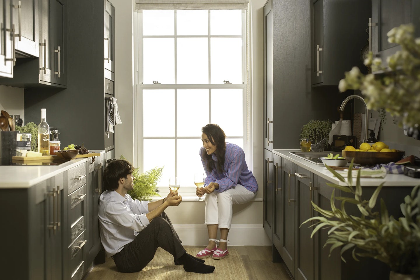 Two people in a modern kitchen sitting near a large window. One person is seated on the floor holding a drink, while the other sits on the windowsill with a glass in hand. The kitchen features dark cabinets, a wooden floor, and countertops with items including a bottle of wine, glasses, and a bowl of fruit. Green plants are visible near the sink and window.