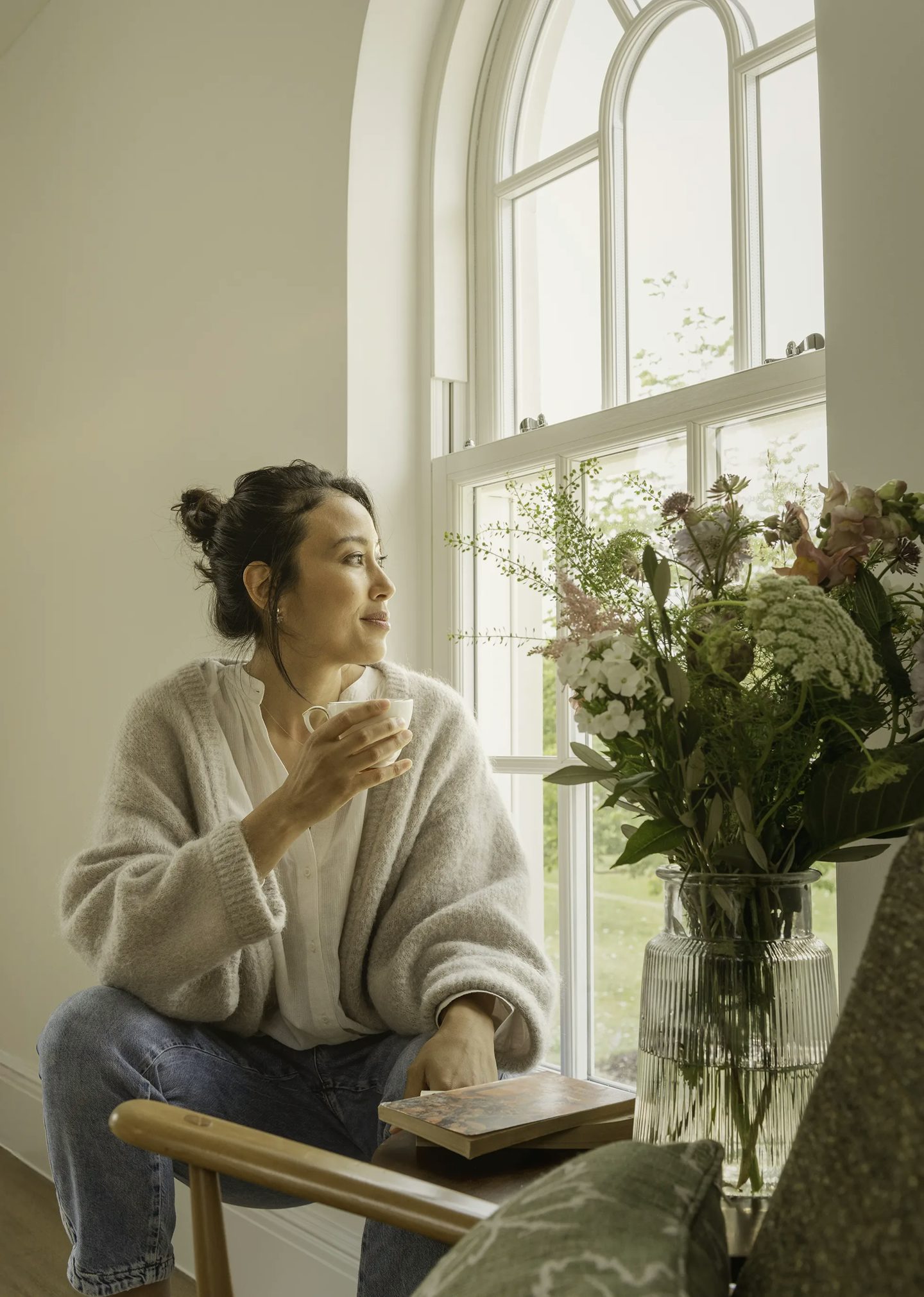 Person sitting on a wooden chair near a tall arched window, holding a cup in one hand and resting the other on a book placed on their lap. A clear glass vase filled with a bouquet of flowers is on a nearby surface, and soft natural light fills the room.