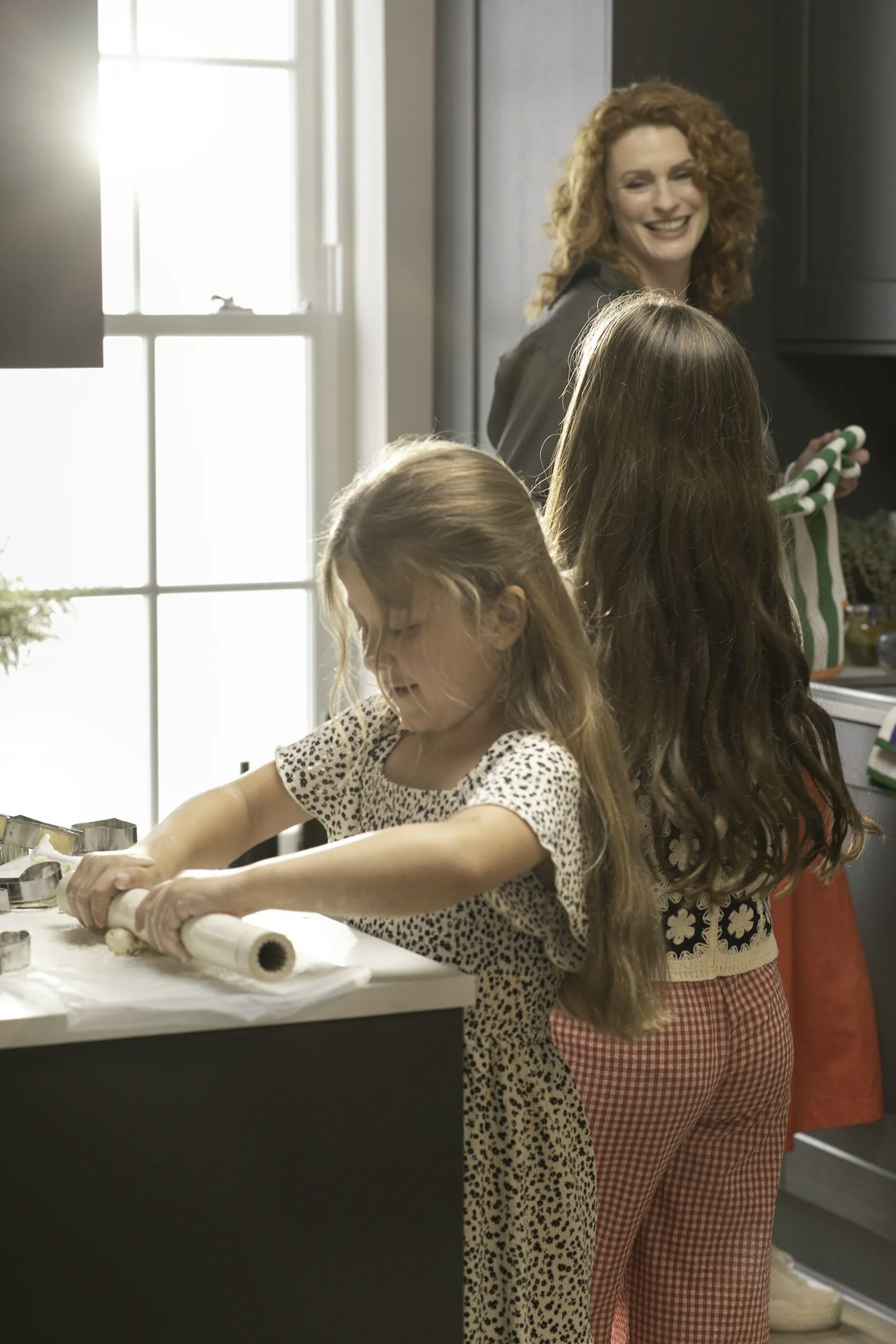 Two children standing at a kitchen counter, one rolling out dough with a rolling pin. Behind them, an adult is near the sink holding a striped bag. The kitchen has dark cabinets, a bright window letting in natural light, and various items on the counter.