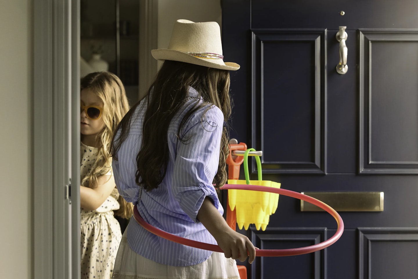 Person wearing a light blue shirt and straw hat holding a pink hula hoop near a dark blue front door. A set of colorful plastic toys, including buckets and spades, hangs on the door. Another person in a light-coloured dress is partially visible in the background inside the house.