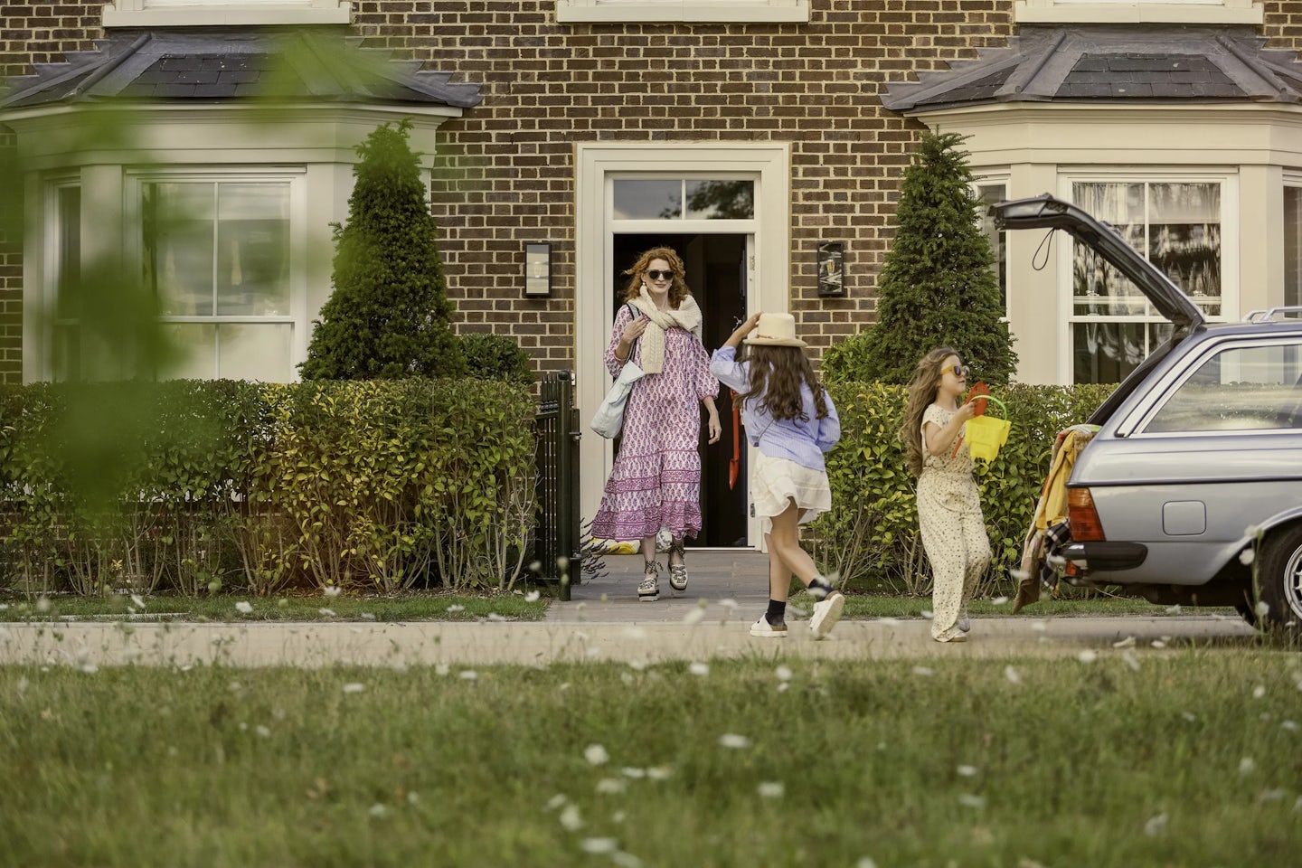 Three people outside a brick house with a cream-colored door and large windows. One person is standing in the doorway holding bags, while two others are near an open car boot on the driveway, carrying colorful buckets and toys. The front garden has neatly trimmed hedges and tall conical shrubs.