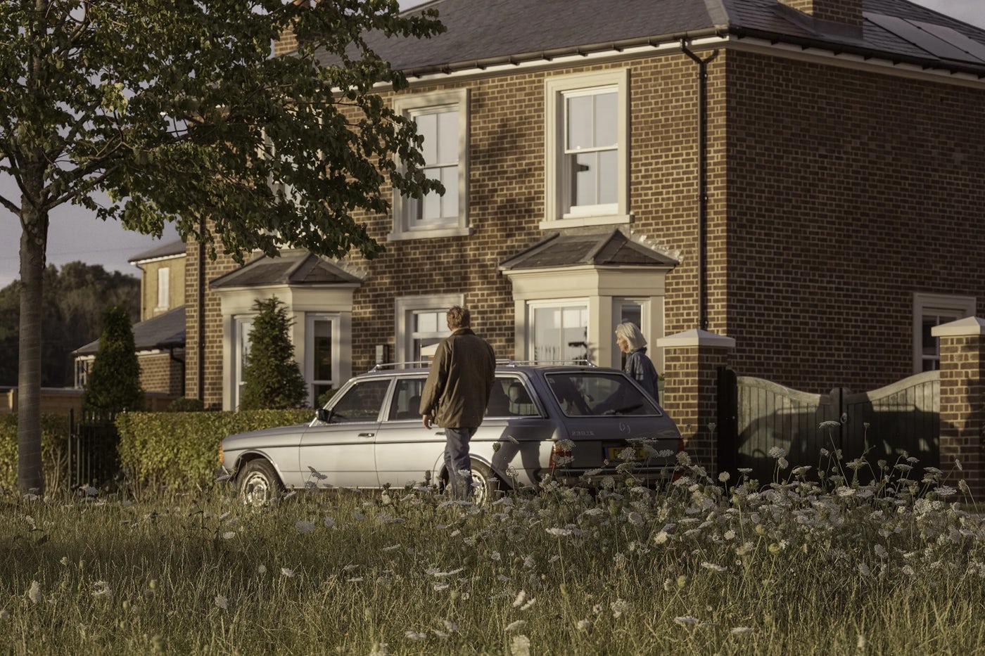 Two people standing near a silver car parked in front of a modern brick house with white-framed windows and a cream-colored door. The house has a small front garden with trimmed hedges and conical shrubs, and a tree is visible on the left side of the image. The foreground shows a grassy area with wildflowers.