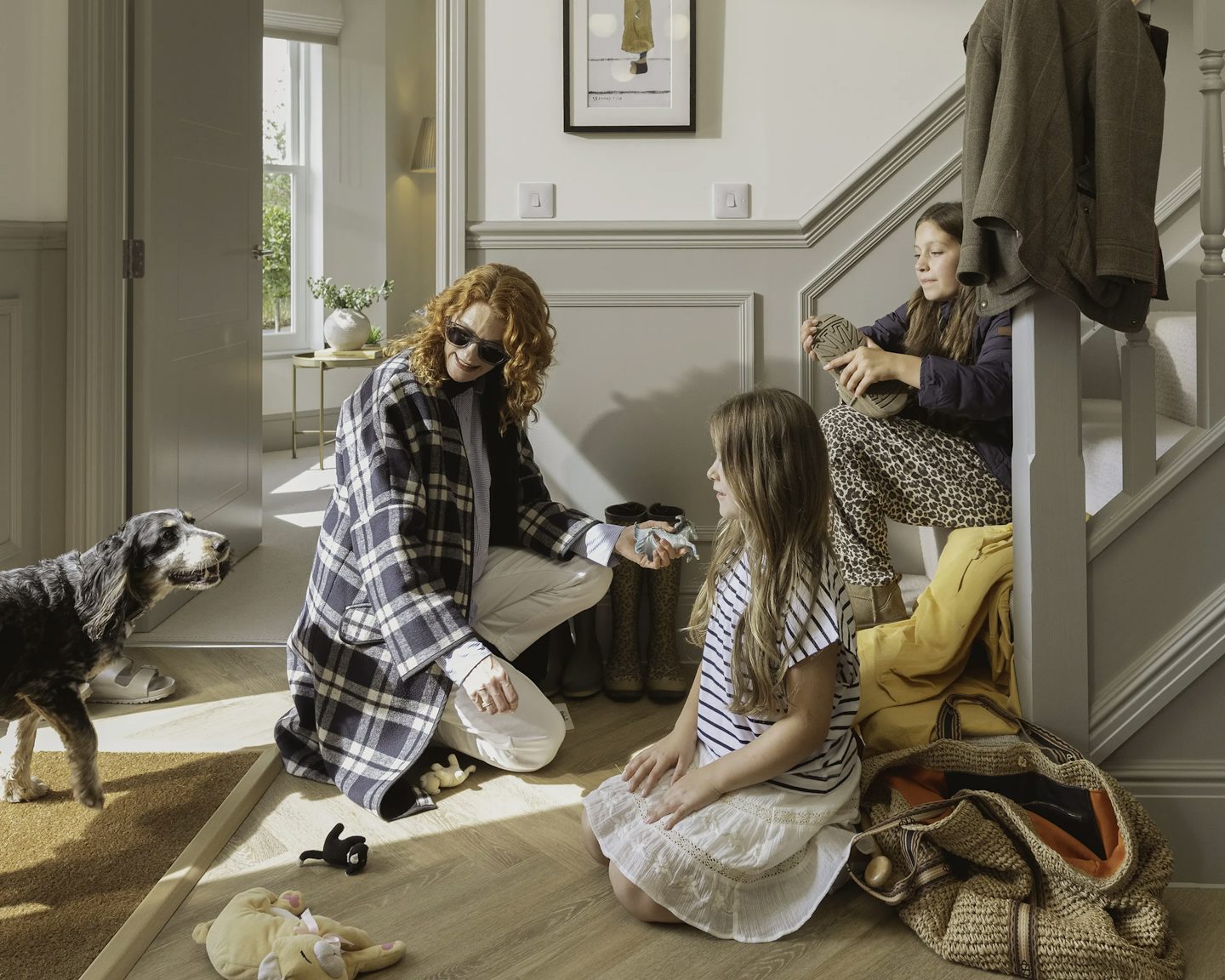 Three people in a hallway near a staircase, with a dog standing on a doormat. One person is kneeling on the wooden floor next to a pair of boots, another is sitting on the bottom step holding an item, and the third is seated on the floor. There are woven bags and soft toys scattered on the floor, a coat draped over the banister, and a framed artwork on the wall.