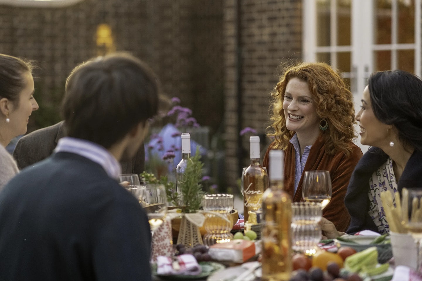 Outdoor dining scene with several people seated around a table covered in food and drinks. The table includes bottles of rosé wine, wine glasses, plates of fruit, breadsticks, and decorative greenery. In the background is a brick building with large windows and purple flowers.