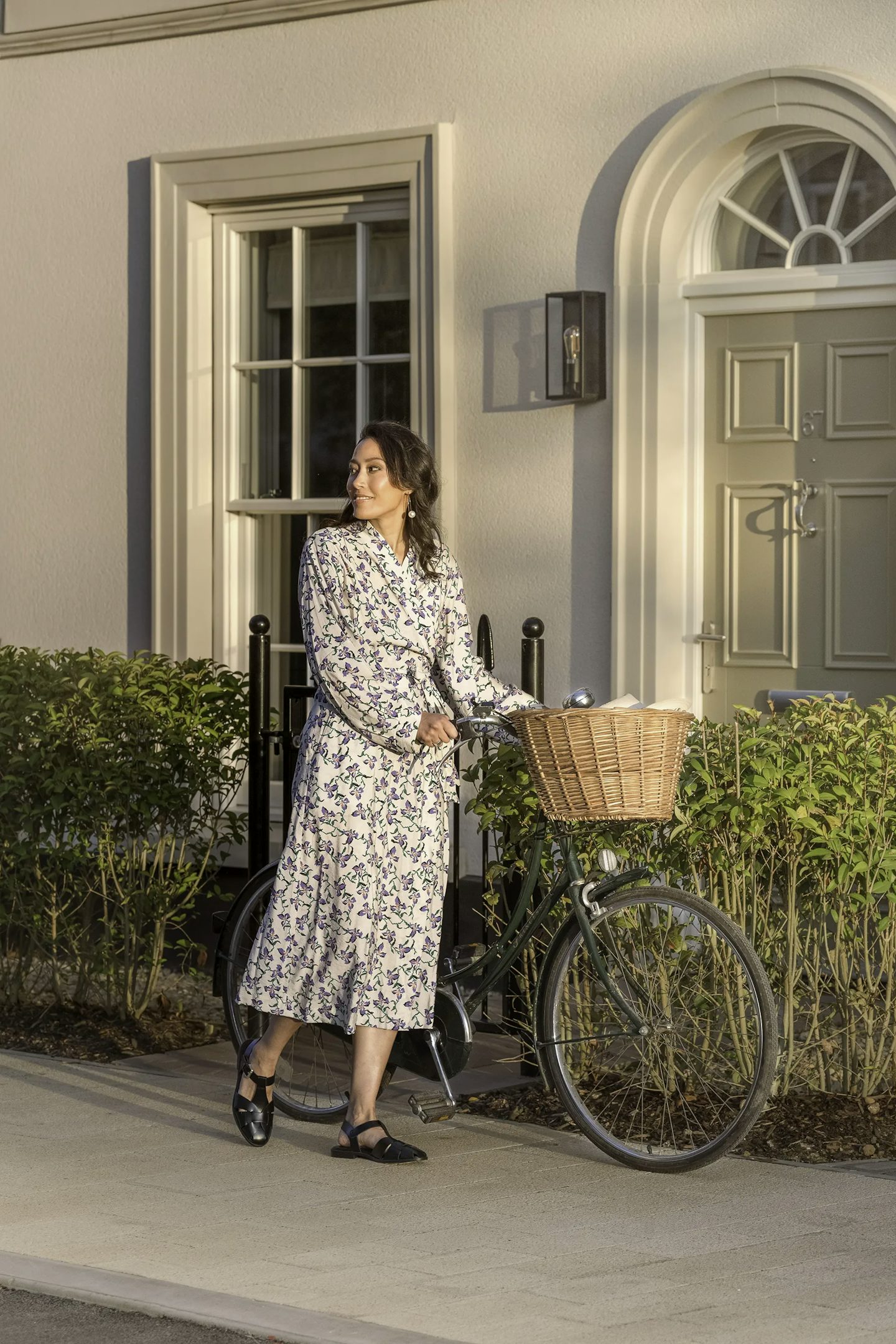 Person standing on a paved path outside a cream-colored house, holding a black bicycle with a wicker basket attached to the handlebars. The house features an arched doorway, a panelled front door, and a large window with white trim. Low green hedges line the front of the property.