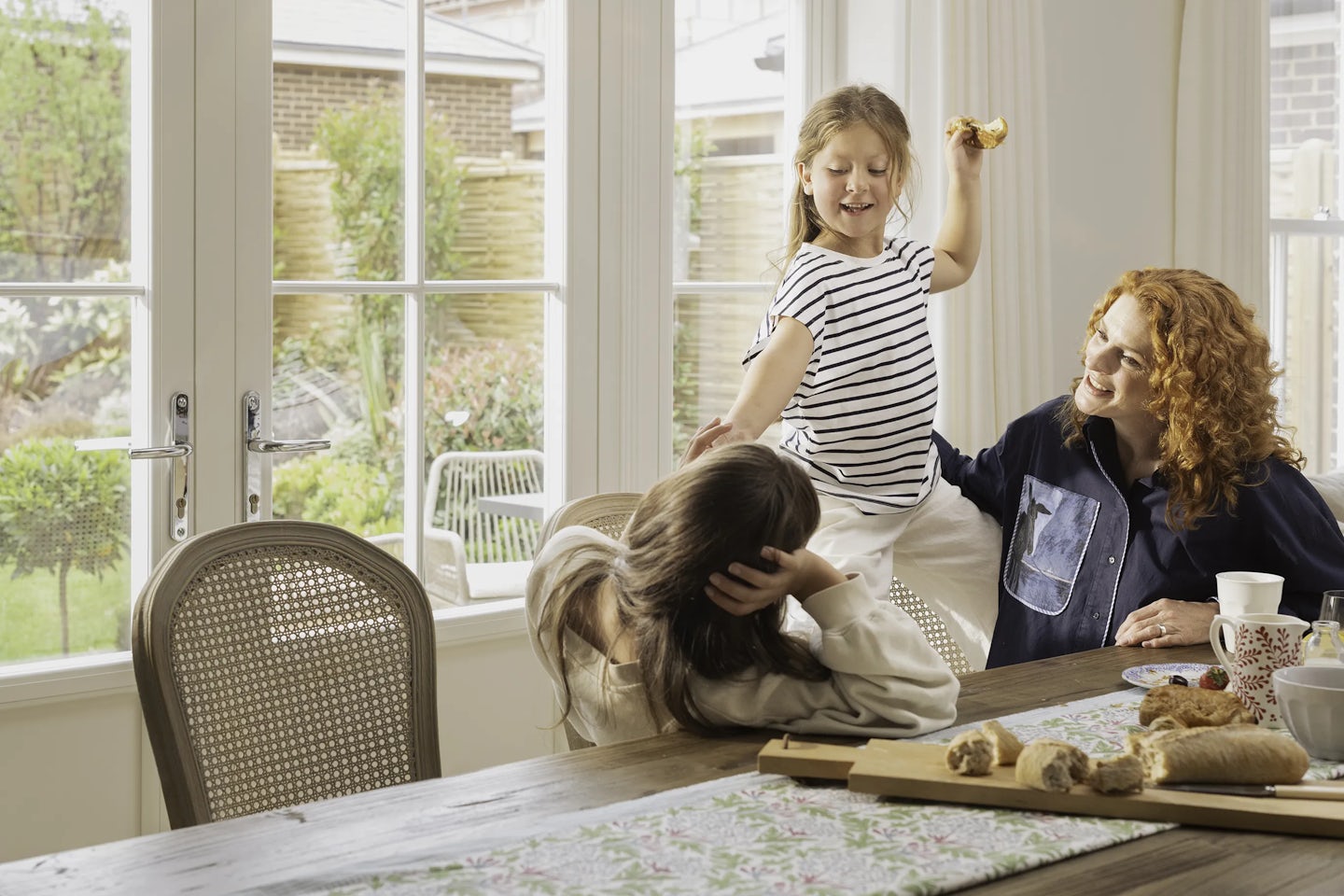 Three people sitting at a wooden dining table in a bright room with large glass doors opening to a garden. One person is leaning on the table, another is seated with a cup, and a child is standing on a chair holding a piece of bread. The table is set with pastries, cups, and a floral-patterned runner.