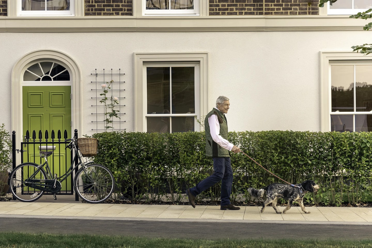 Person walking a black-and-white dog along a paved path in front of a cream-colored house with a bright green door and large windows. A black bicycle with a wicker basket is parked against a black metal fence near the doorway. Neatly trimmed hedges line the front of the property.