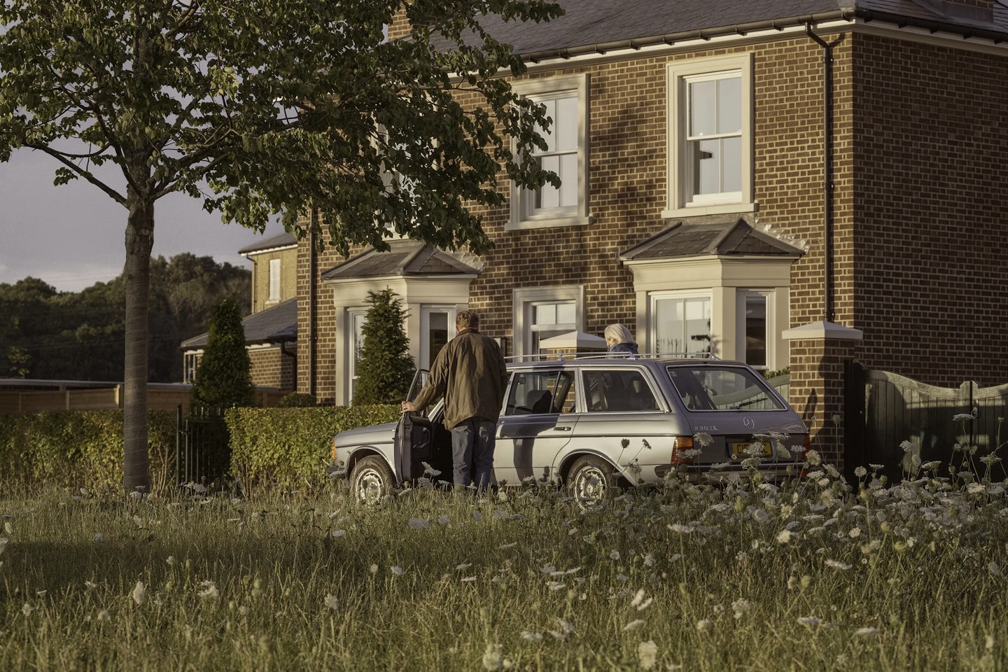 Two people standing beside a silver estate car parked on a driveway in front of a modern brick house with large white-framed windows and a cream-colored door. The car’s boot is open, and one person is holding a black bag. A tree and tall grass with wildflowers are in the foreground, and the house is surrounded by neatly trimmed hedges.