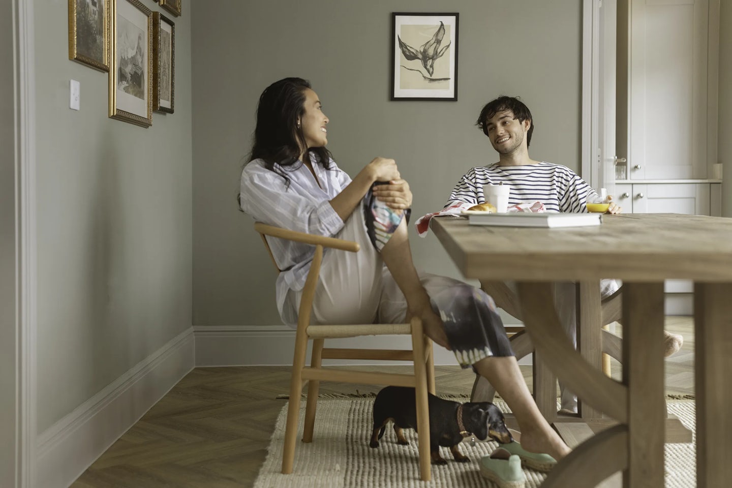 Two people sitting at a wooden dining table in a modern room with light-colored walls and framed artwork. One person is seated on a wooden chair with legs crossed, while the other sits at the table with a cup and a plate in front. A small black dachshund stands on a woven rug near the chair. The background includes a doorway and additional wall art.