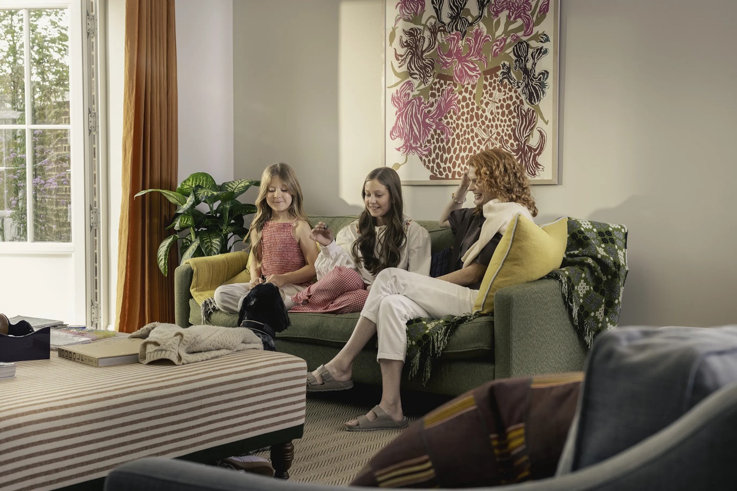 Three people sitting on a green sofa in a living room. The sofa has patterned and solid-colored cushions, and a green throw draped over the armrest. A black-and-white dog is sitting on the floor near the sofa. Behind the group is a large colorful abstract artwork on the wall, and to the left is a tall green plant next to a glass door with orange curtains. In the foreground, there is a striped ottoman with books and a blanket.