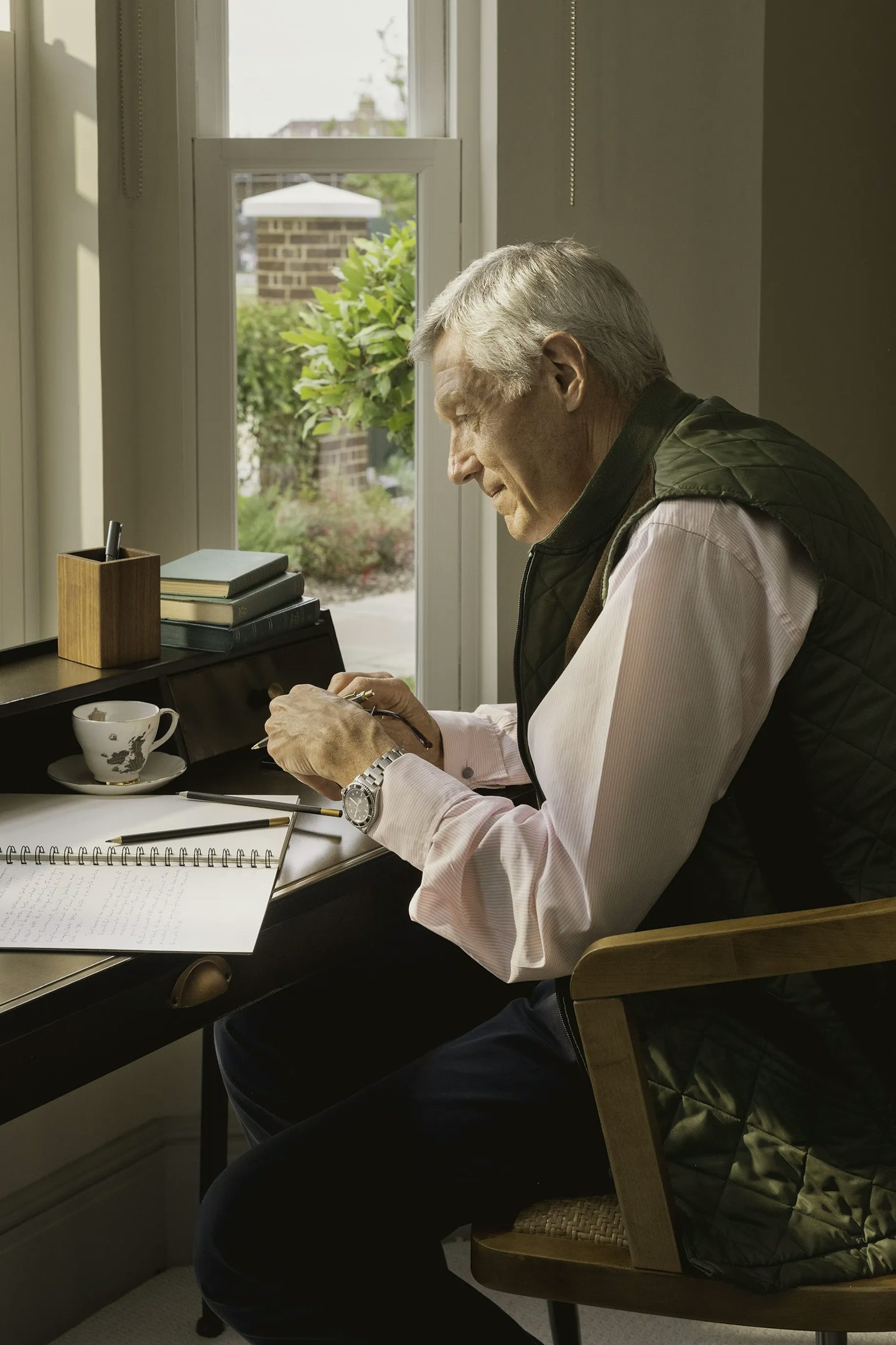 Person seated at a wooden desk near a large window, writing in a notebook. The desk has a cup and saucer, a wooden pen holder, and a small stack of books. Outside the window, greenery and part of a brick wall are visible.
