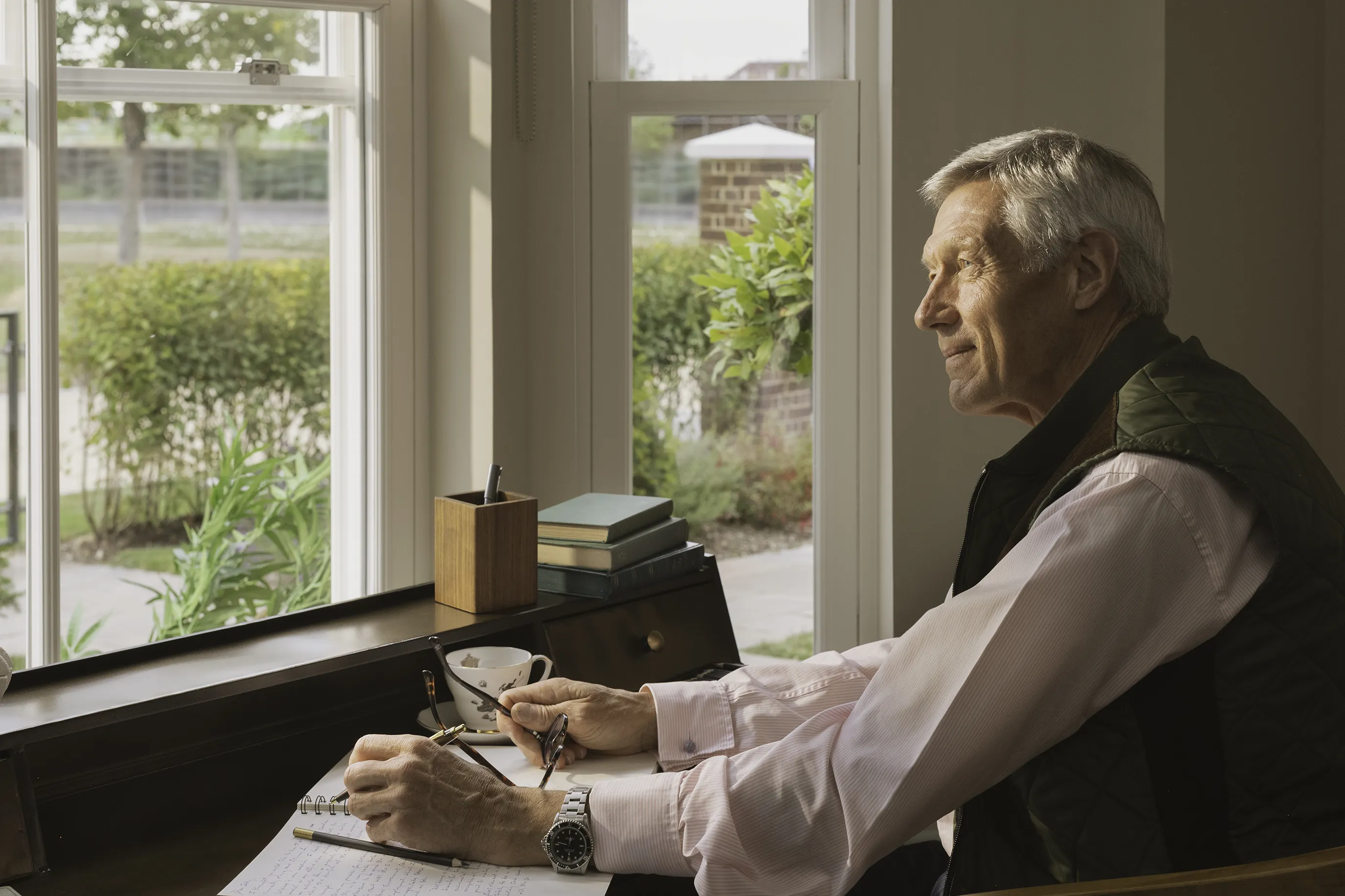 Person seated at a wooden desk near a large window, writing in a notebook with a pen. A pair of glasses rests on the desk alongside a cup of coffee, a wooden pen holder, and a small stack of books. Outside the window, greenery and a brick pillar are visible.
