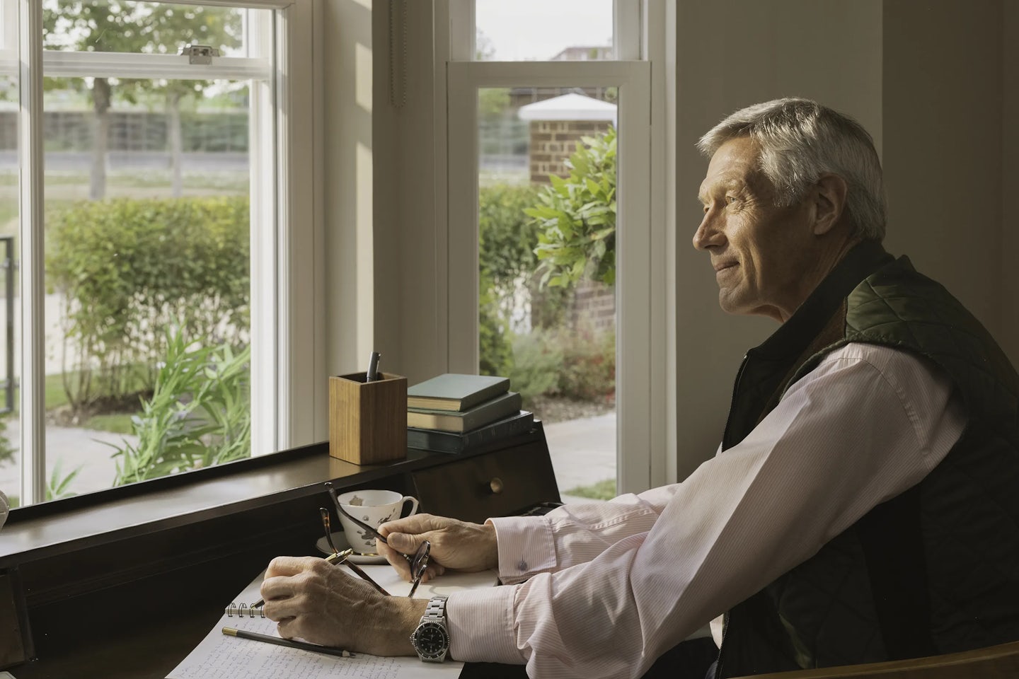 Person seated at a wooden desk near a large window, writing in a notebook with a pen. A pair of glasses rests on the desk alongside a cup of coffee, a wooden pen holder, and a small stack of books. Outside the window, greenery and a brick pillar are visible.
