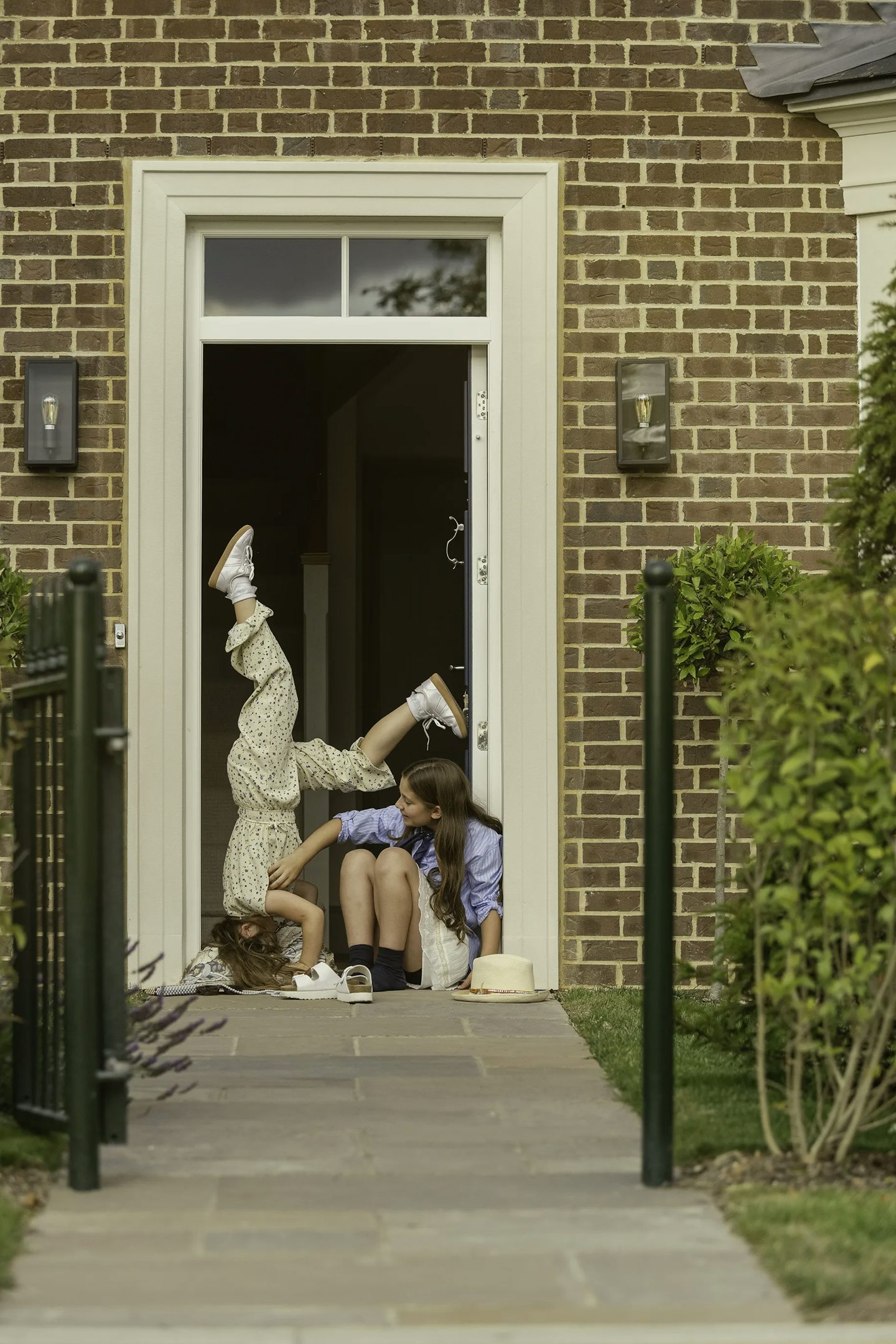 Two children sitting in the doorway of a brick house, with one child performing a handstand while the other sits beside them. A straw hat is placed on the ground near the doorway, and the entrance is framed by cream-colored trim. A paved path leads up to the door, bordered by green hedges and a black metal gate.