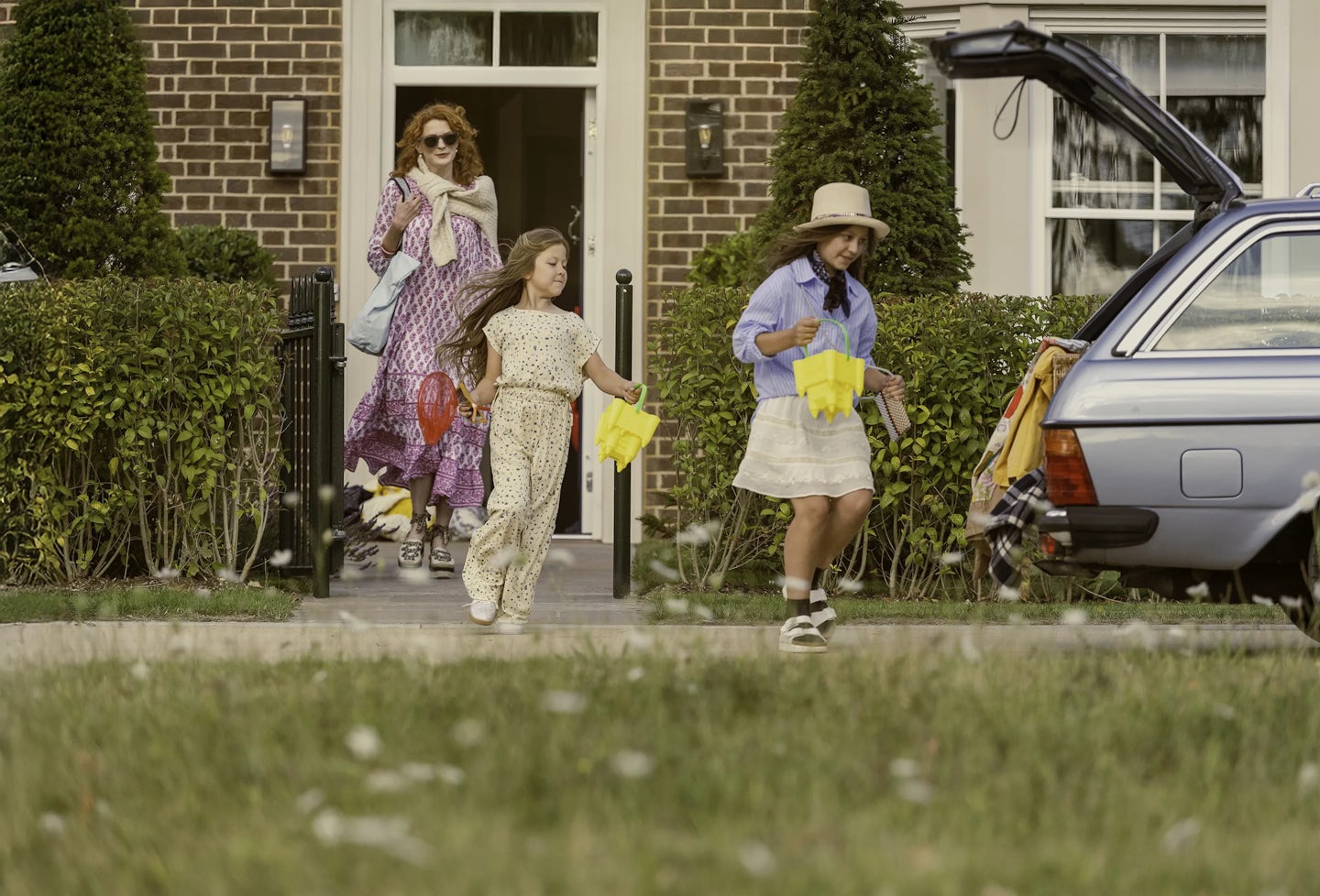 Three people outside a brick house with a cream-colored door, walking toward a silver car with its boot open. The car is parked on a driveway, and colorful buckets and toys are visible near the boot. The individuals are carrying bags and beach toys. The front garden has neatly trimmed hedges and tall conical shrubs, and the foreground shows a grassy area with wildflowers.
