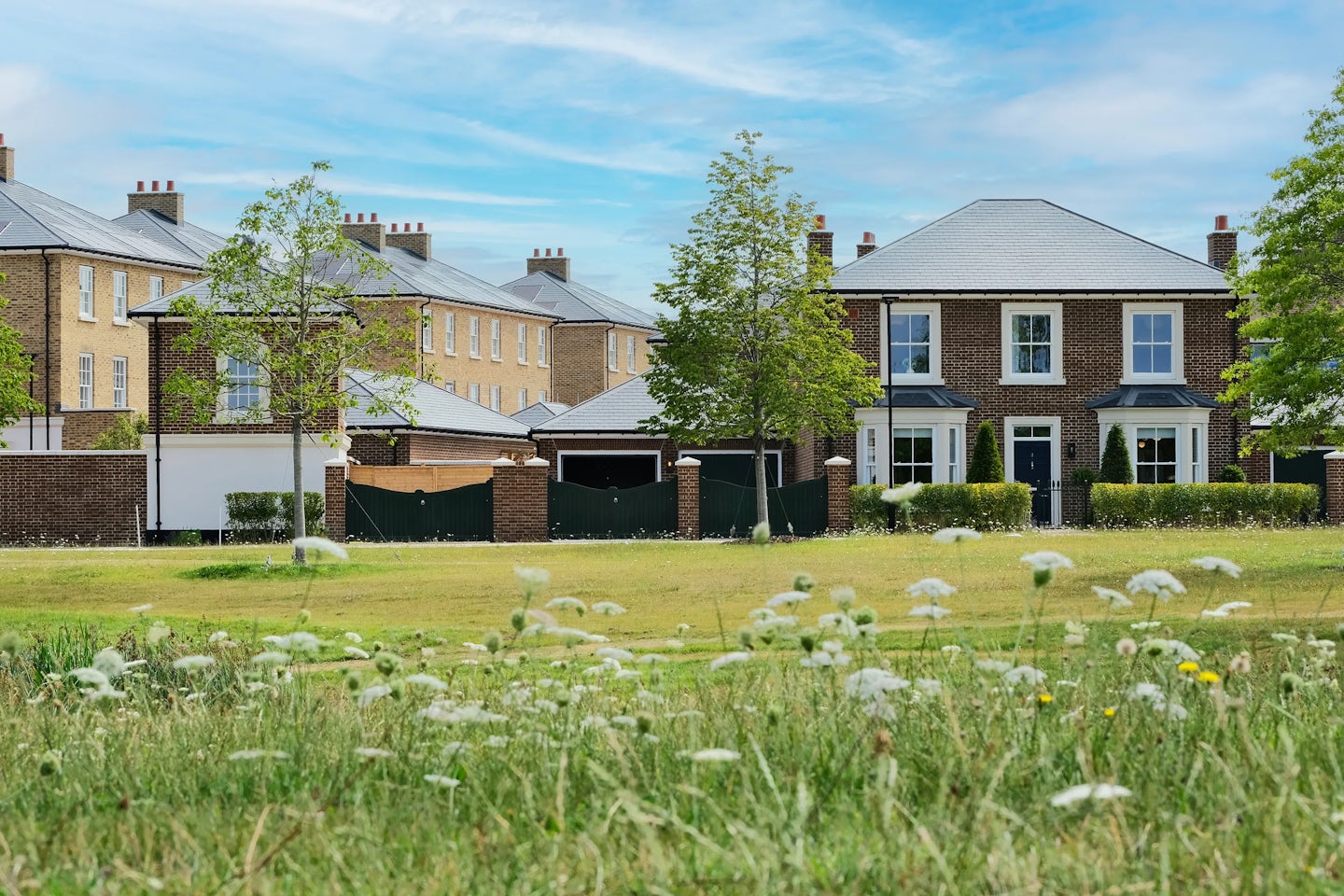 View of a residential development featuring multiple modern brick houses with pitched slate roofs and white-framed windows. The foreground shows a grassy area with wildflowers and young trees, while the background includes several homes arranged along a street, partially enclosed by black fencing and brick pillars. The sky is bright blue with scattered clouds.