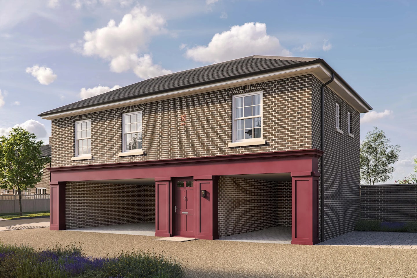 Two-story brick building with a pitched roof and white trim. The upper level has three white-framed sash windows, while the lower level features three open carport bays and a central door with a letterbox, all framed in dark red. The building sits on a gravel driveway with a small landscaped area in the foreground, and the sky is partly cloudy with bright sunlight.