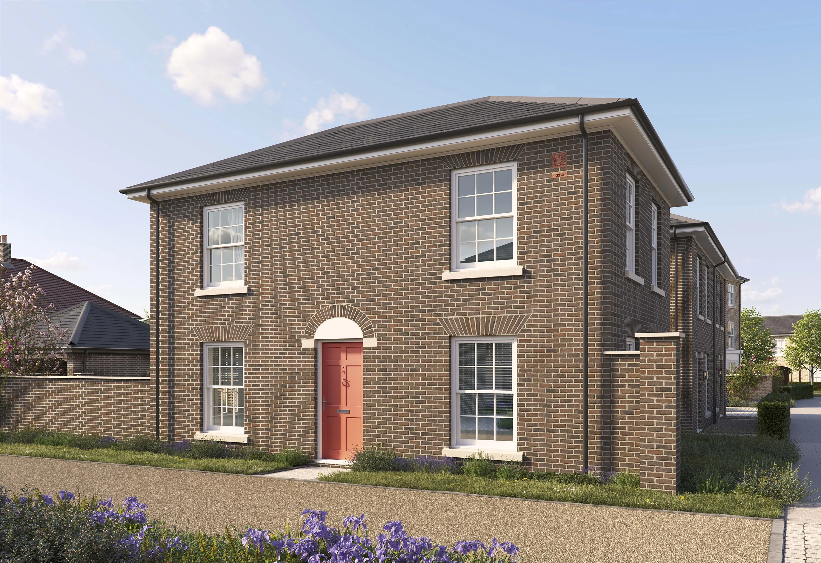 Two-story brick house with a pitched roof and white trim. The front features a red door framed by an arched brick detail, two large white-framed windows on the ground floor, and two similar windows on the upper floor. The house sits on a paved path with a small grass verge and purple flowers in the foreground. The sky is bright blue with scattered clouds.