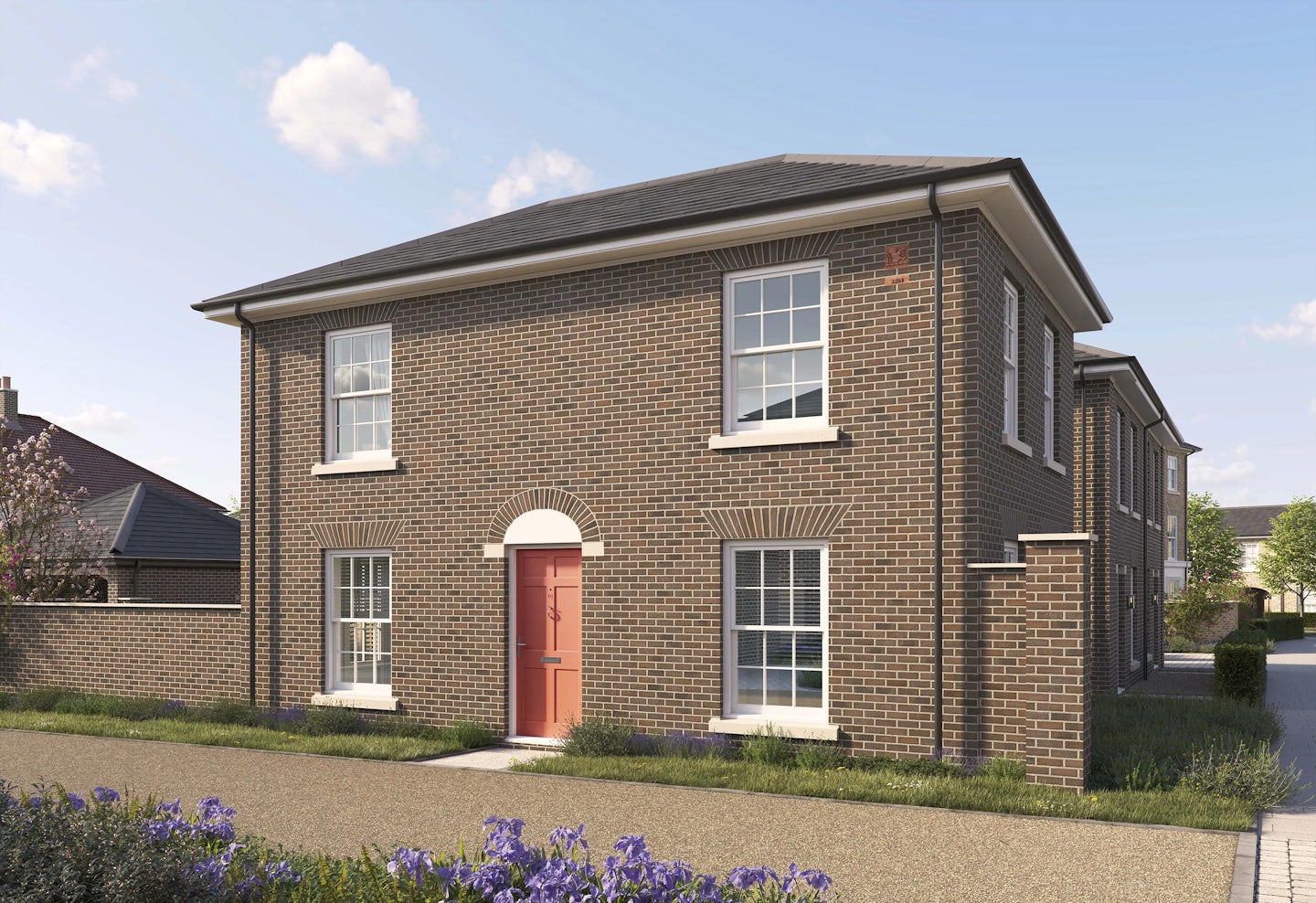 Two-story brick house with a pitched roof and white trim. The front features a red door framed by an arched brick detail, two large white-framed windows on the ground floor, and two similar windows on the upper floor. The house sits on a paved path with a small grass verge and purple flowers in the foreground. The sky is bright blue with scattered clouds.